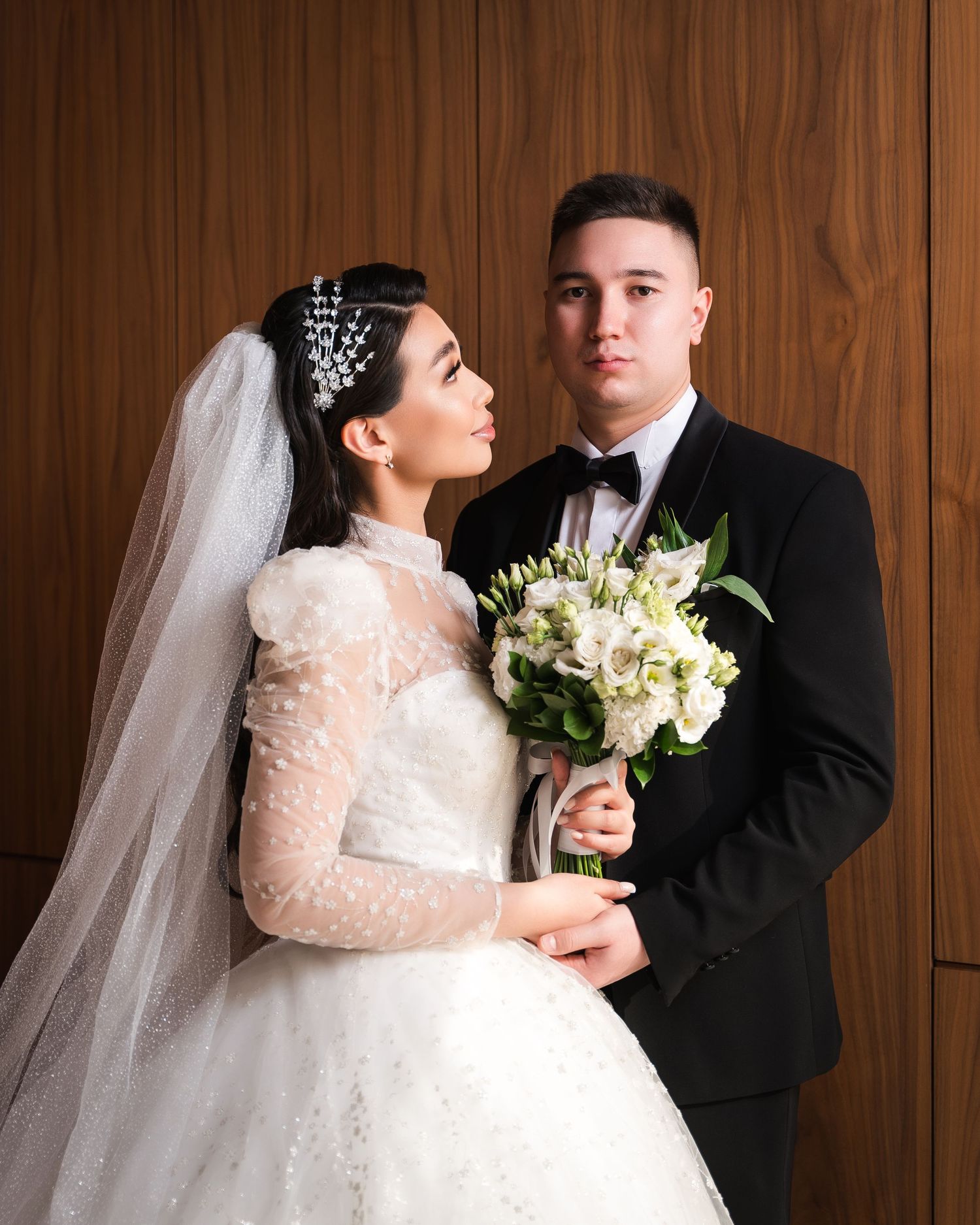Elegant bride in white lace gown with veil and groom in black tuxedo posing together holding a white floral bouquet.
