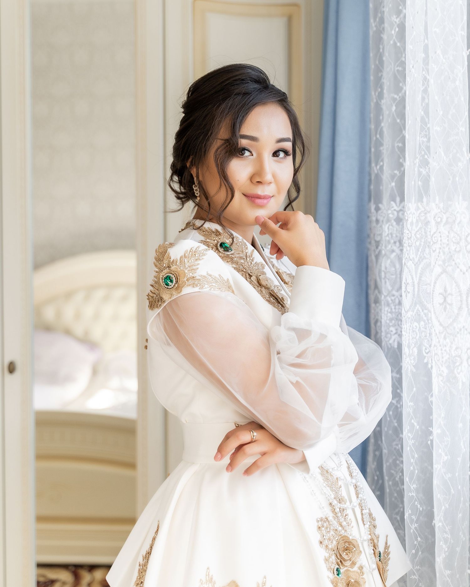 Young woman in elegant white and gold embroidered traditional bridal dress with green gems posing gracefully by a window.
