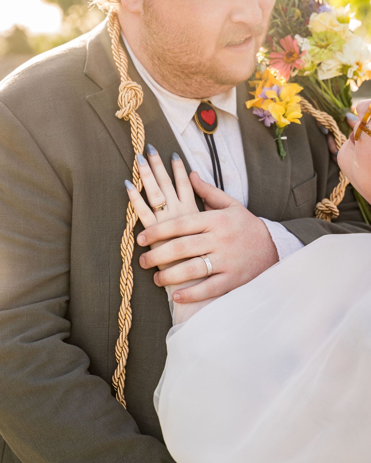 Groom in gray suit with bolo tie and rope detail as bride places hand on his chest holding bouquet.