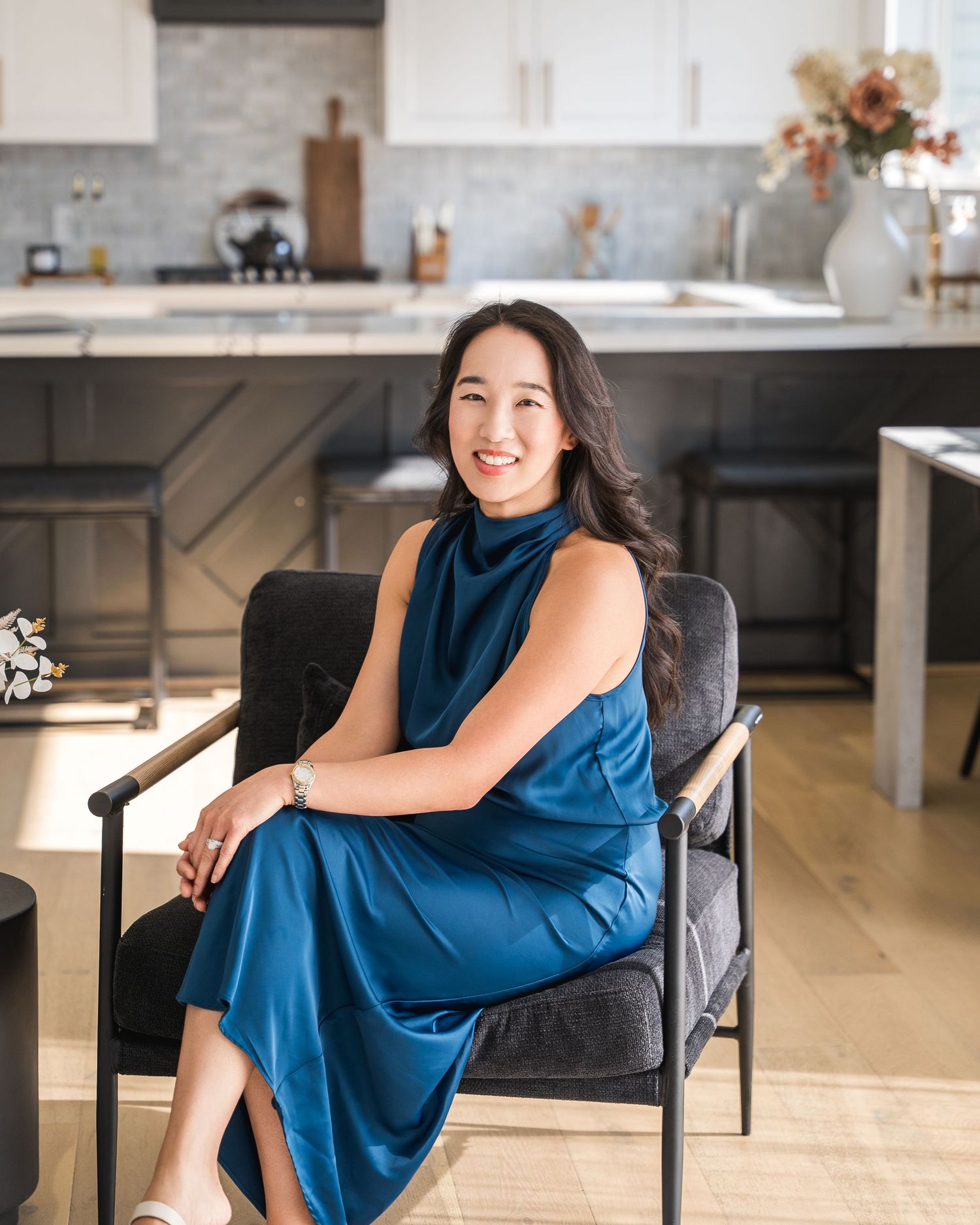 Person in blue dress seated in modern chair against kitchen backdrop.
