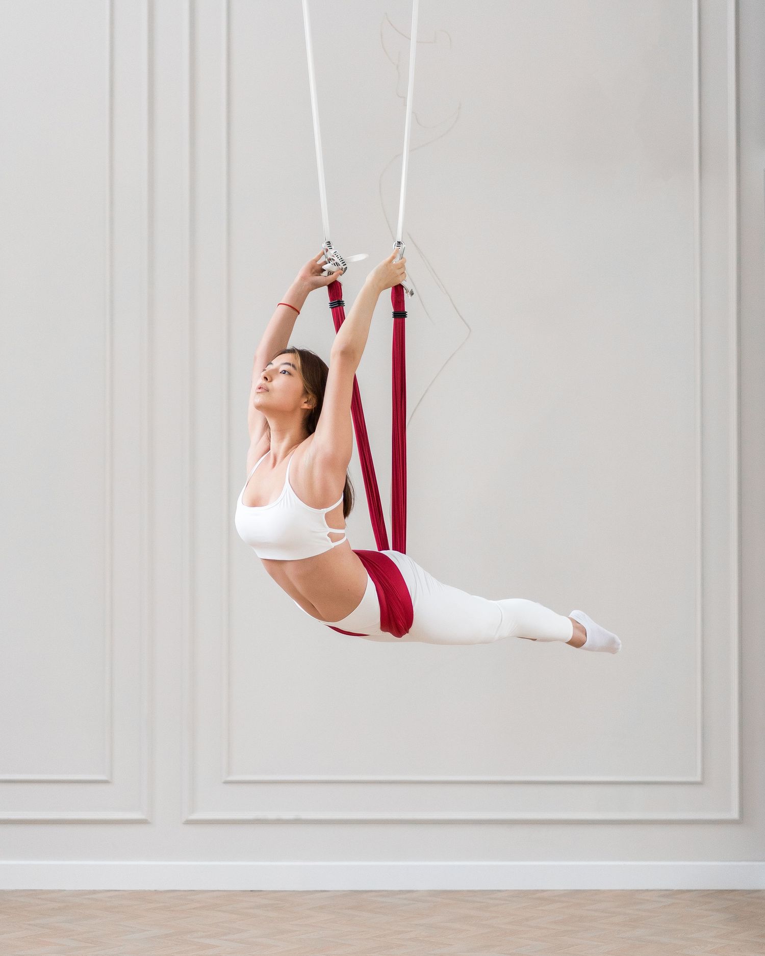 Aerial yoga performer executing suspended poses with red straps against white wall.