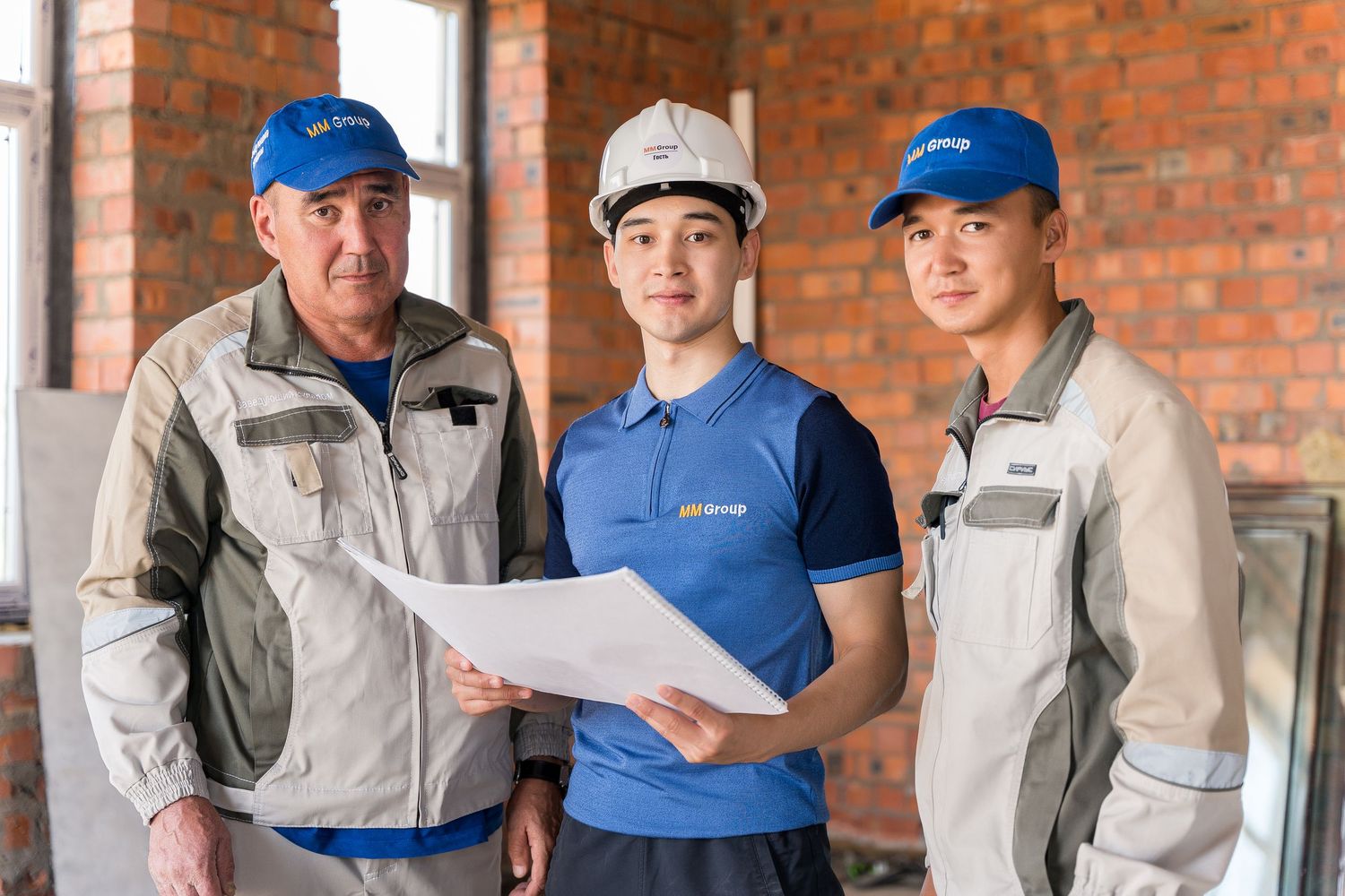 Construction workers in blue uniforms and hard hats reviewing building plans at a brick site.
