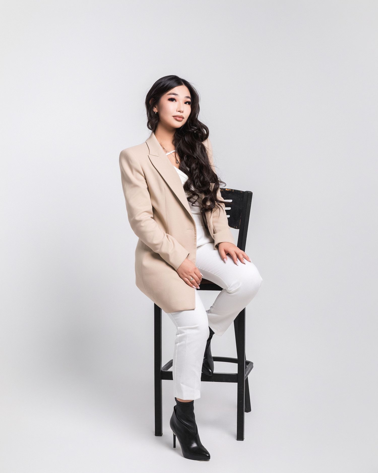 Person in beige blazer and white pants posing on black chair against white background.