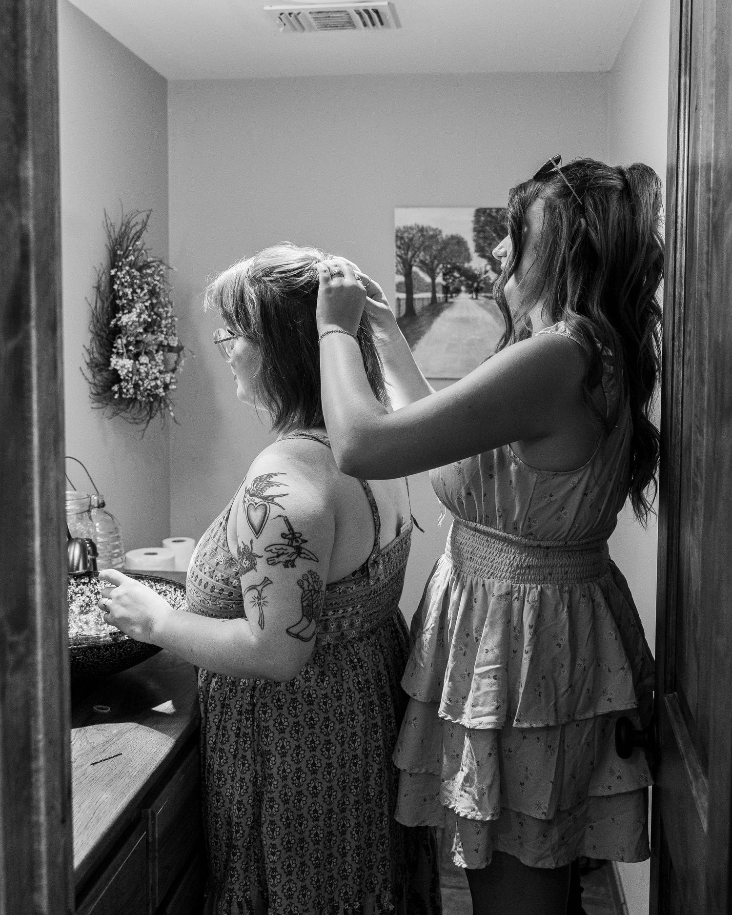 Woman with tattoos gets hair styled by another woman in floral dress in black and white photo.