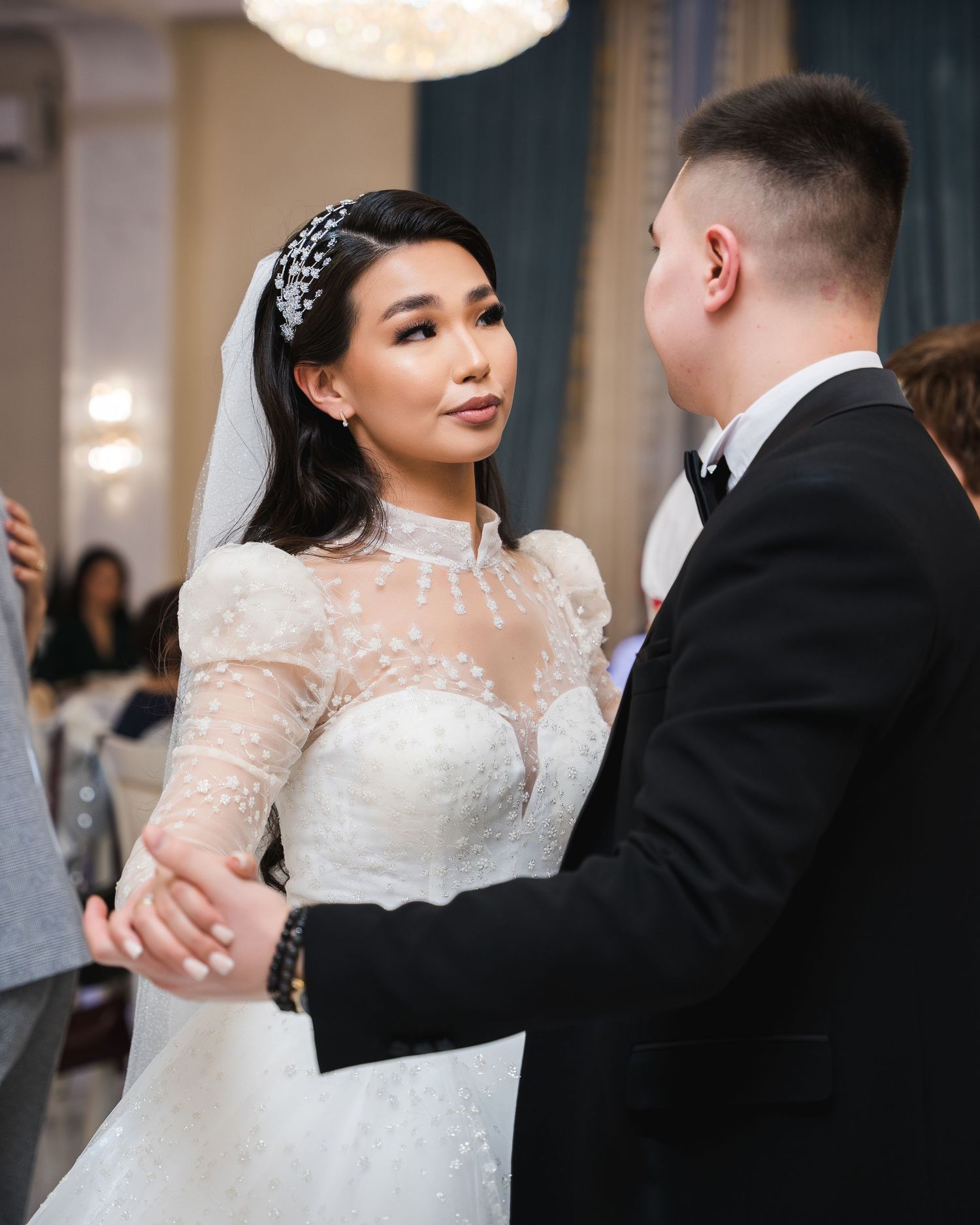 Elegant bride in beaded high-neck wedding gown holds hands with groom in black tuxedo during first dance.