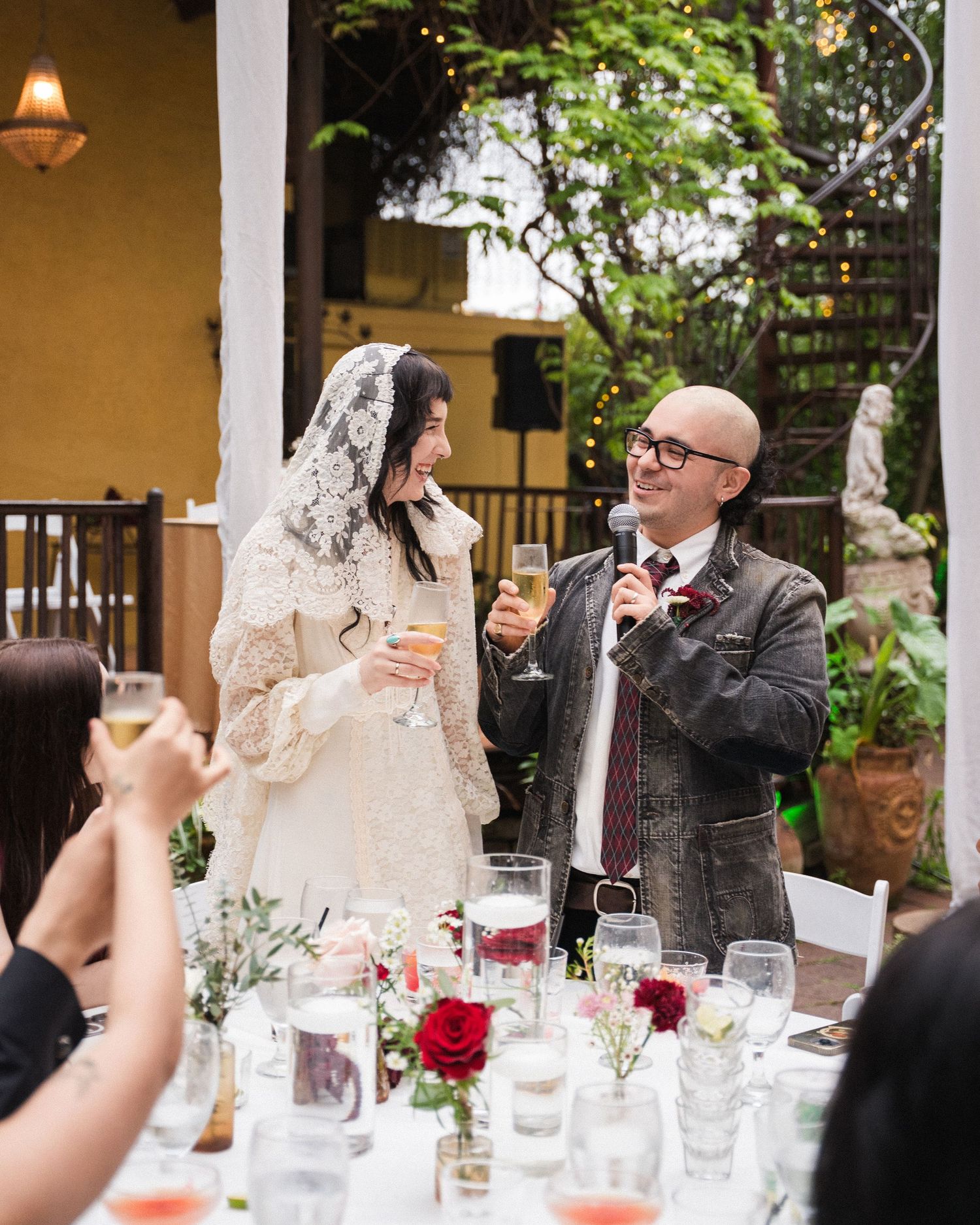 Bride in lace gown and groom in suit share a toast with guests at an elegant outdoor wedding reception.