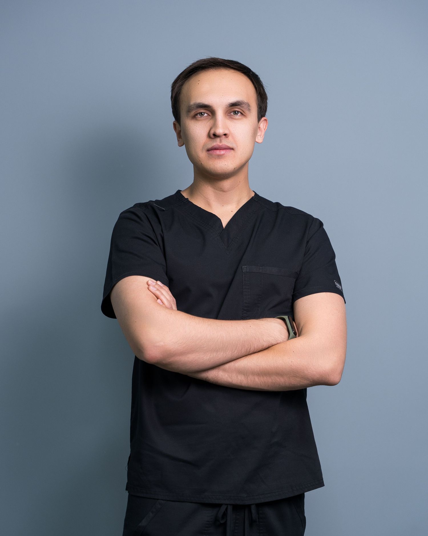 Medical professional in black scrubs posing with crossed arms against blue background.