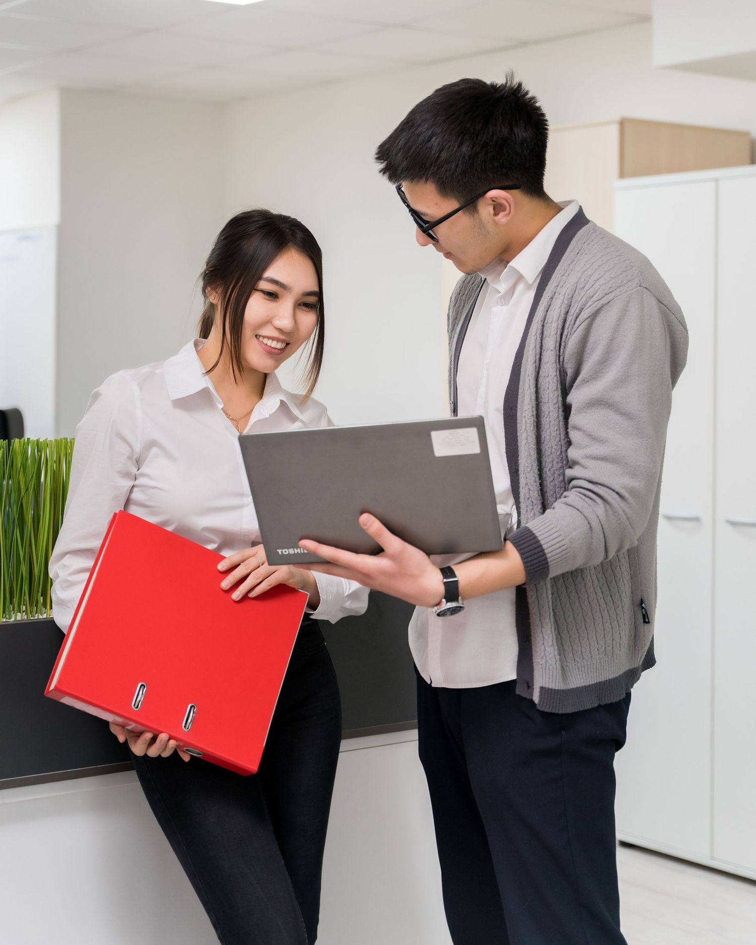 Two colleagues review documents together in a modern office setting, holding folders and discussing work materials.