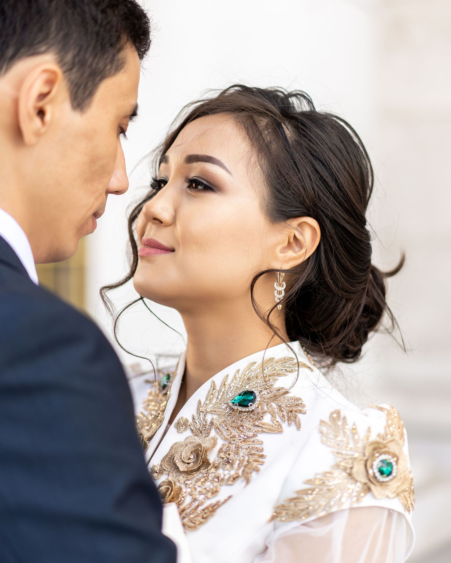 Bride in embroidered white ao dai with gold floral details and emerald earrings gazes at groom during wedding ceremony.