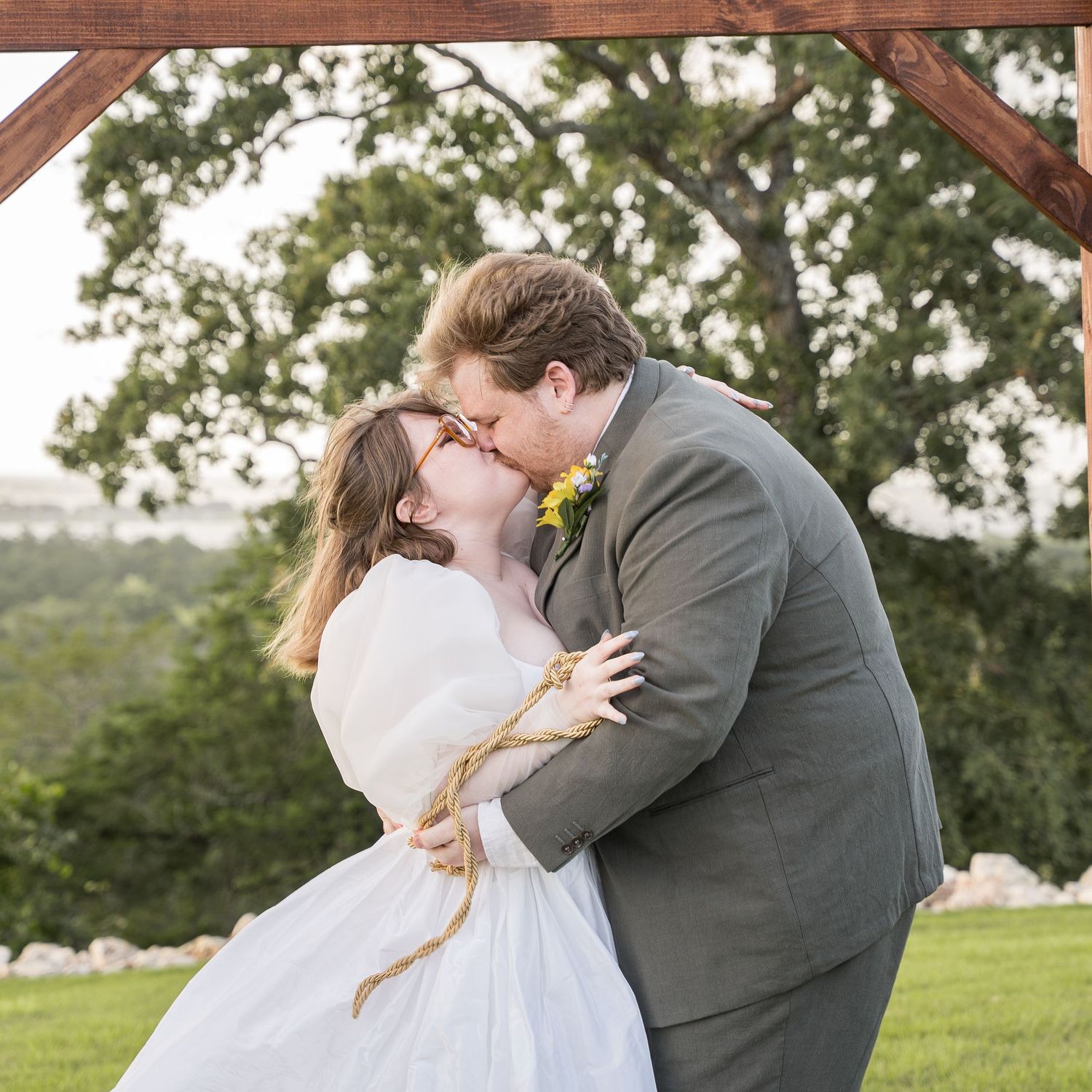 Newlyweds share first kiss under wooden arch surrounded by lush greenery at outdoor wedding.