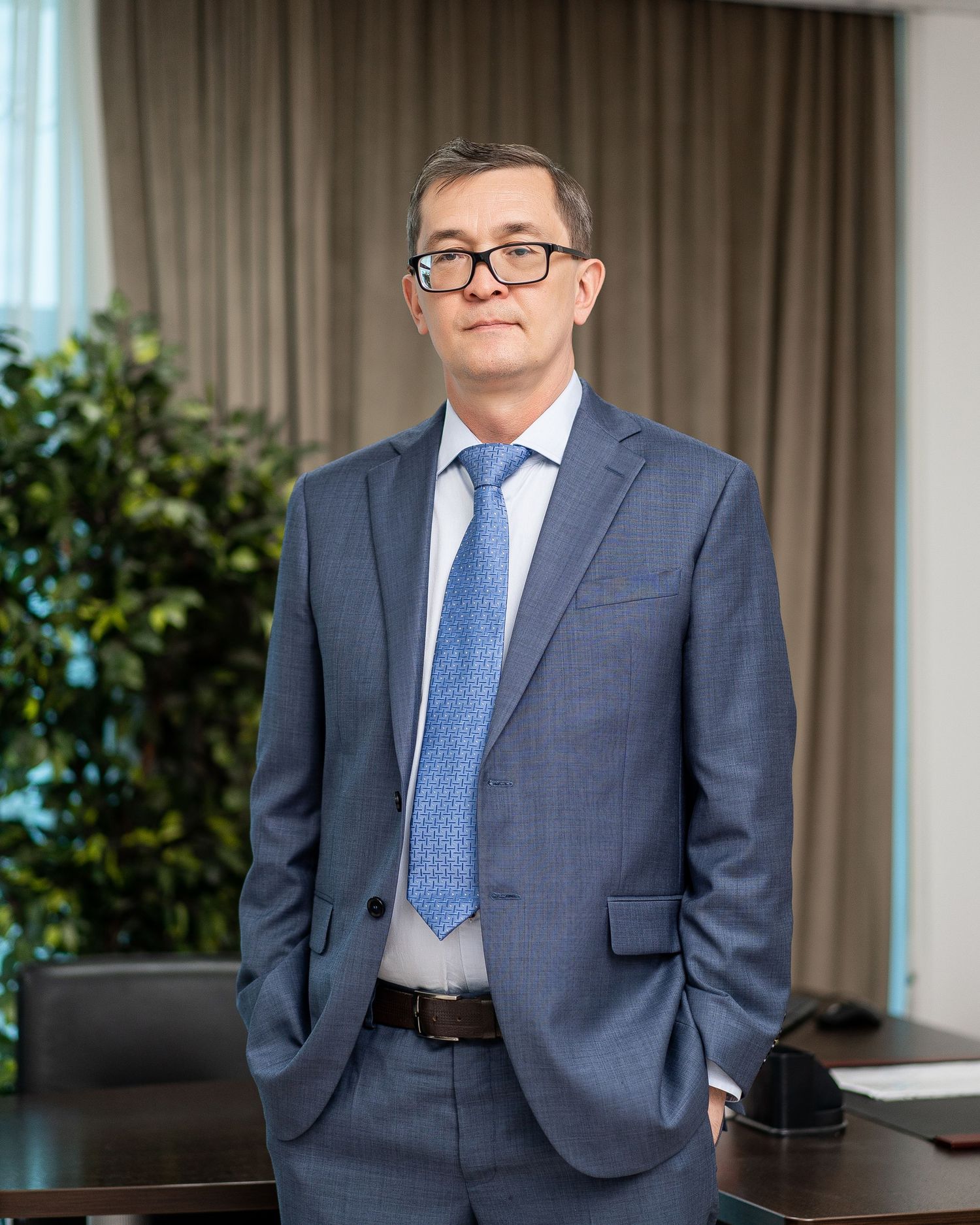 Business professional in navy suit standing against curtained office backdrop.