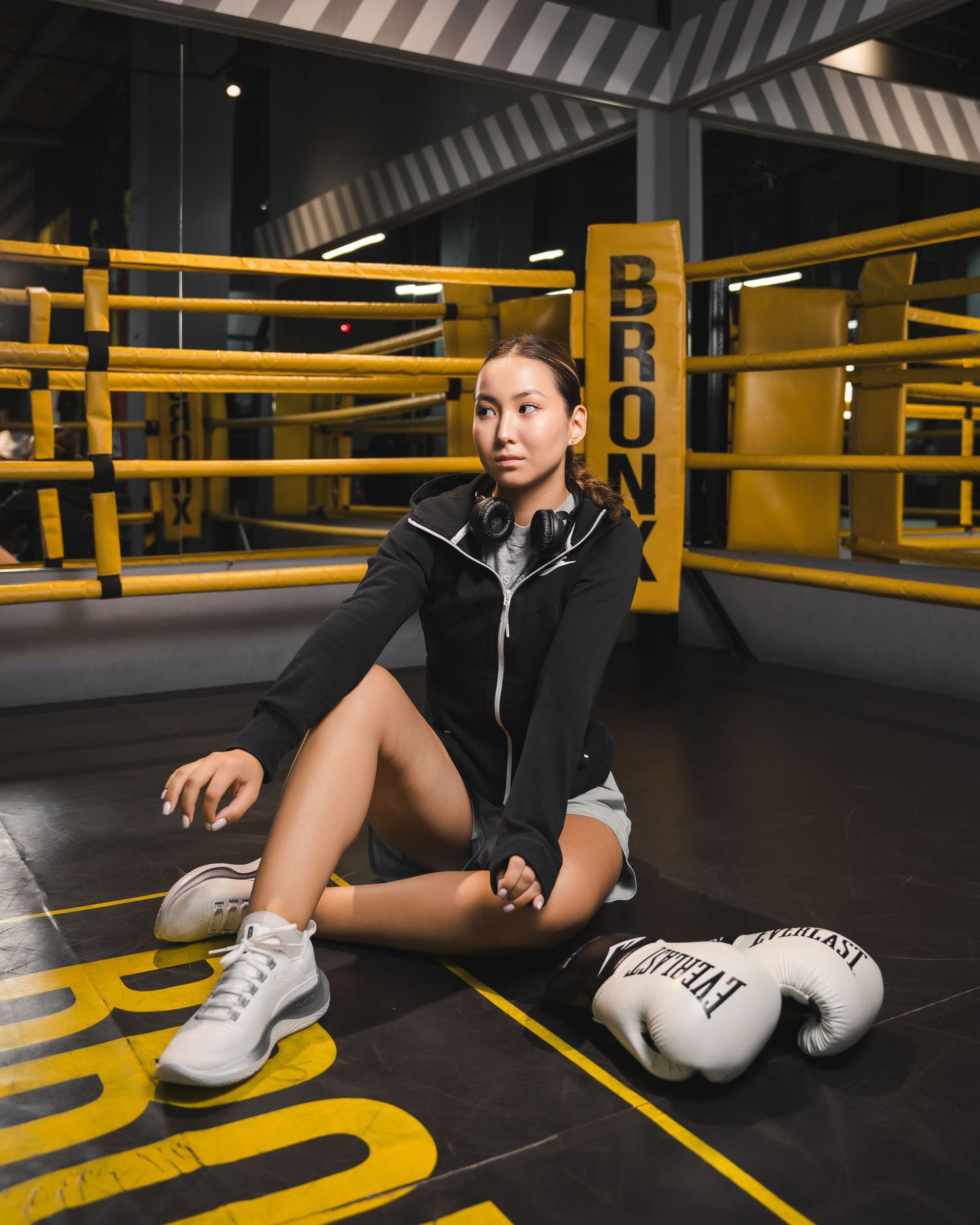 Person in athletic wear stretching on gym floor with boxing gloves nearby.