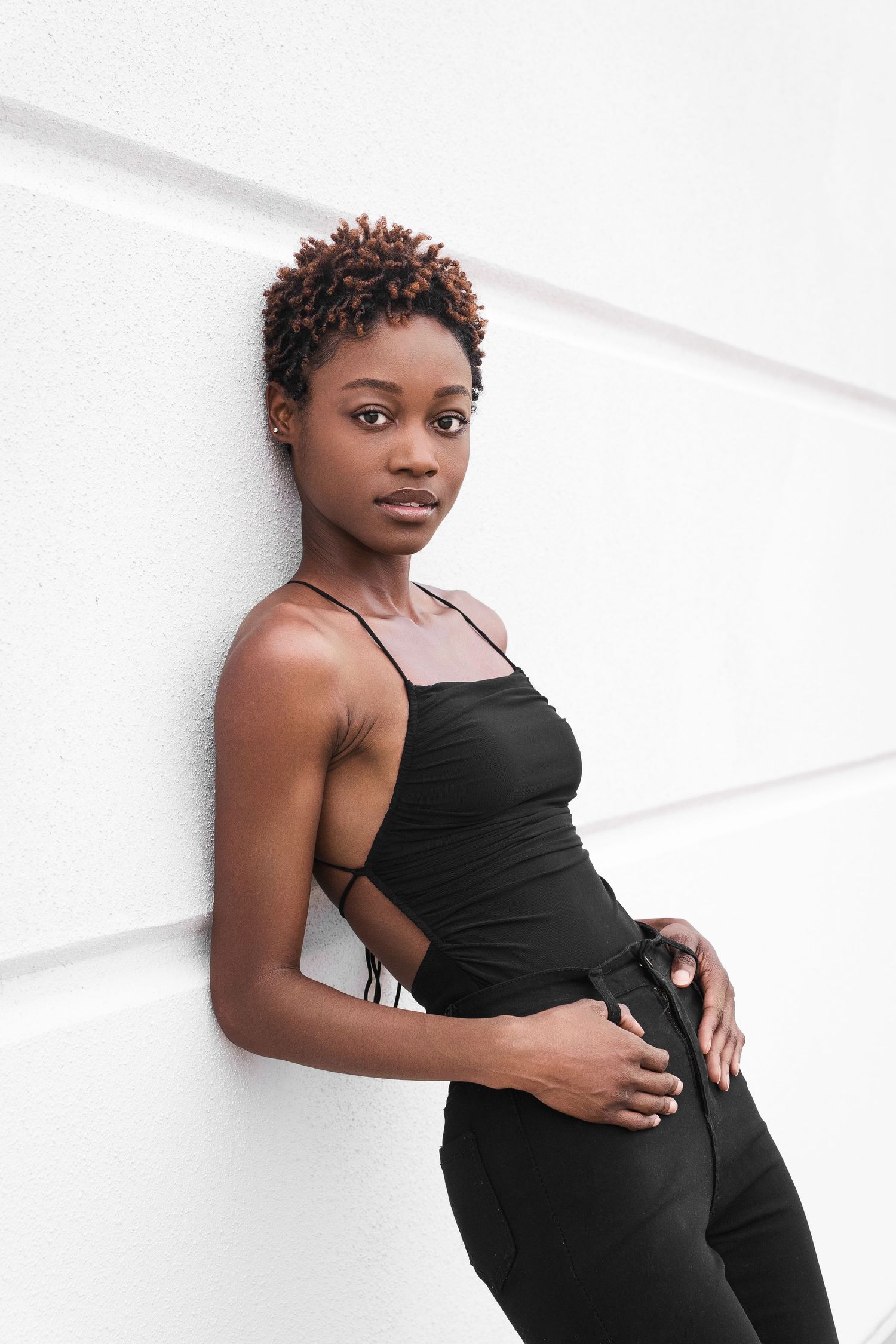 Model in black outfit posing confidently against white wall.