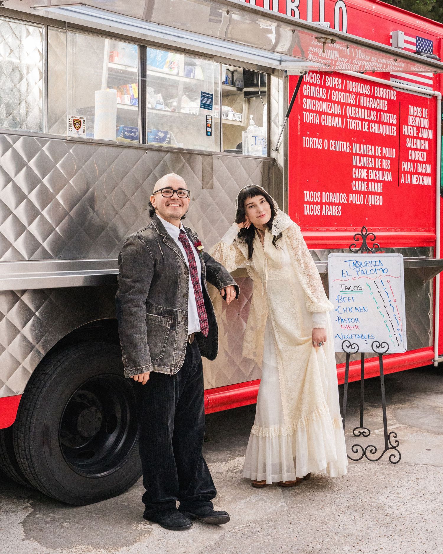 Couple in wedding attire posing in front of a red taco food truck, bride in ivory gown, groom in gray suit.