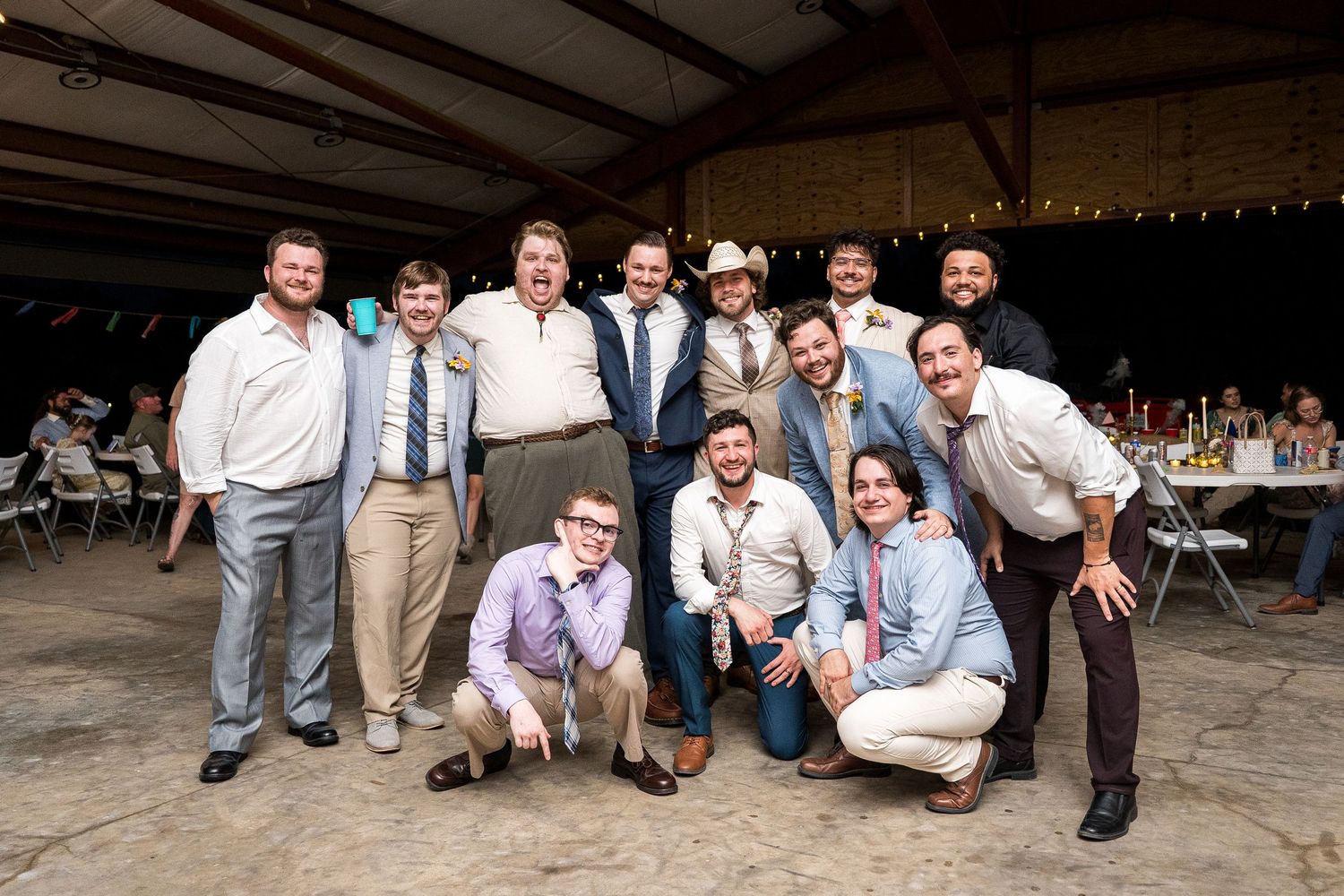 Group of men posing together at a rustic barn wedding reception, smiling for a group photo under string lights.