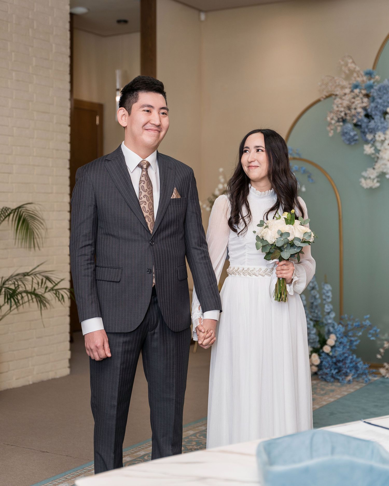 Smiling couple in elegant wedding attire holding hands at a modern ceremony venue decorated with blue floral arrangements.