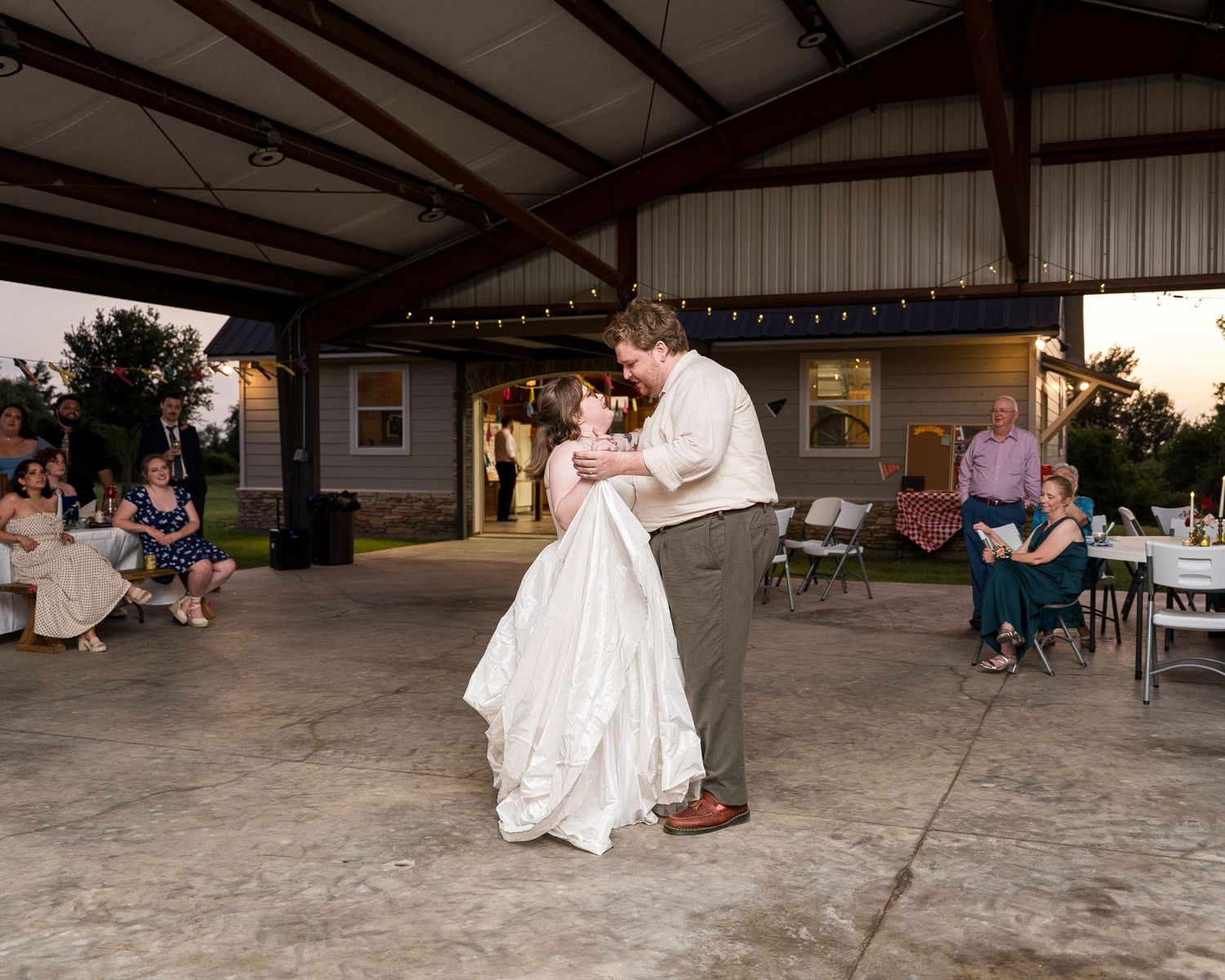 Bride and groom share first dance at outdoor wedding reception under pavilion with string lights at dusk.