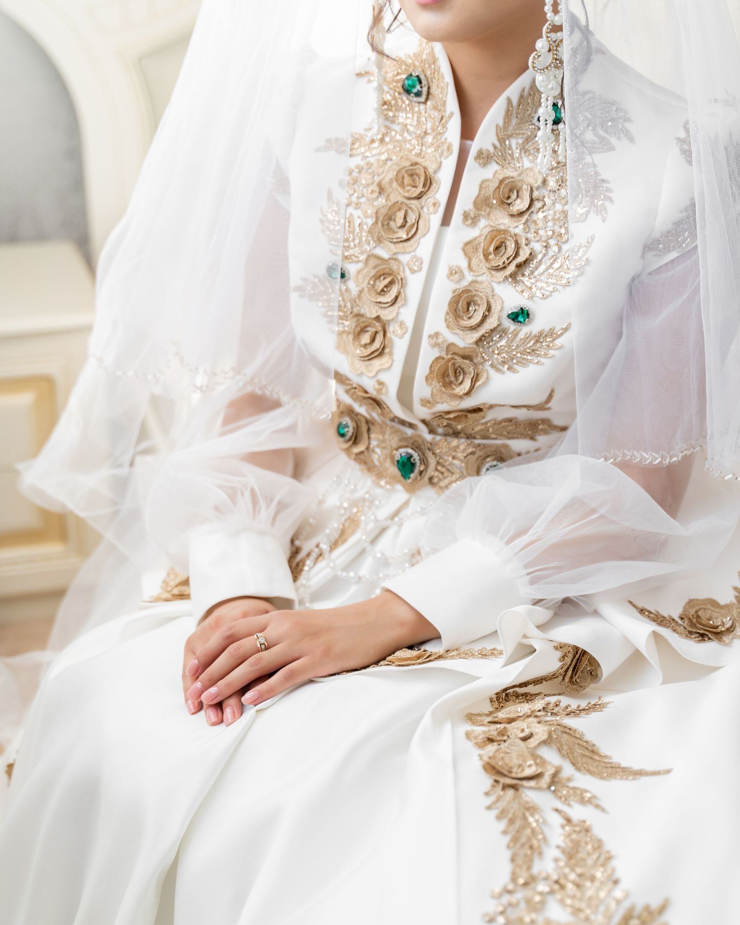 Close-up of white traditional bridal gown with gold rose embroidery, green gems, and sheer veil, worn by seated bride.