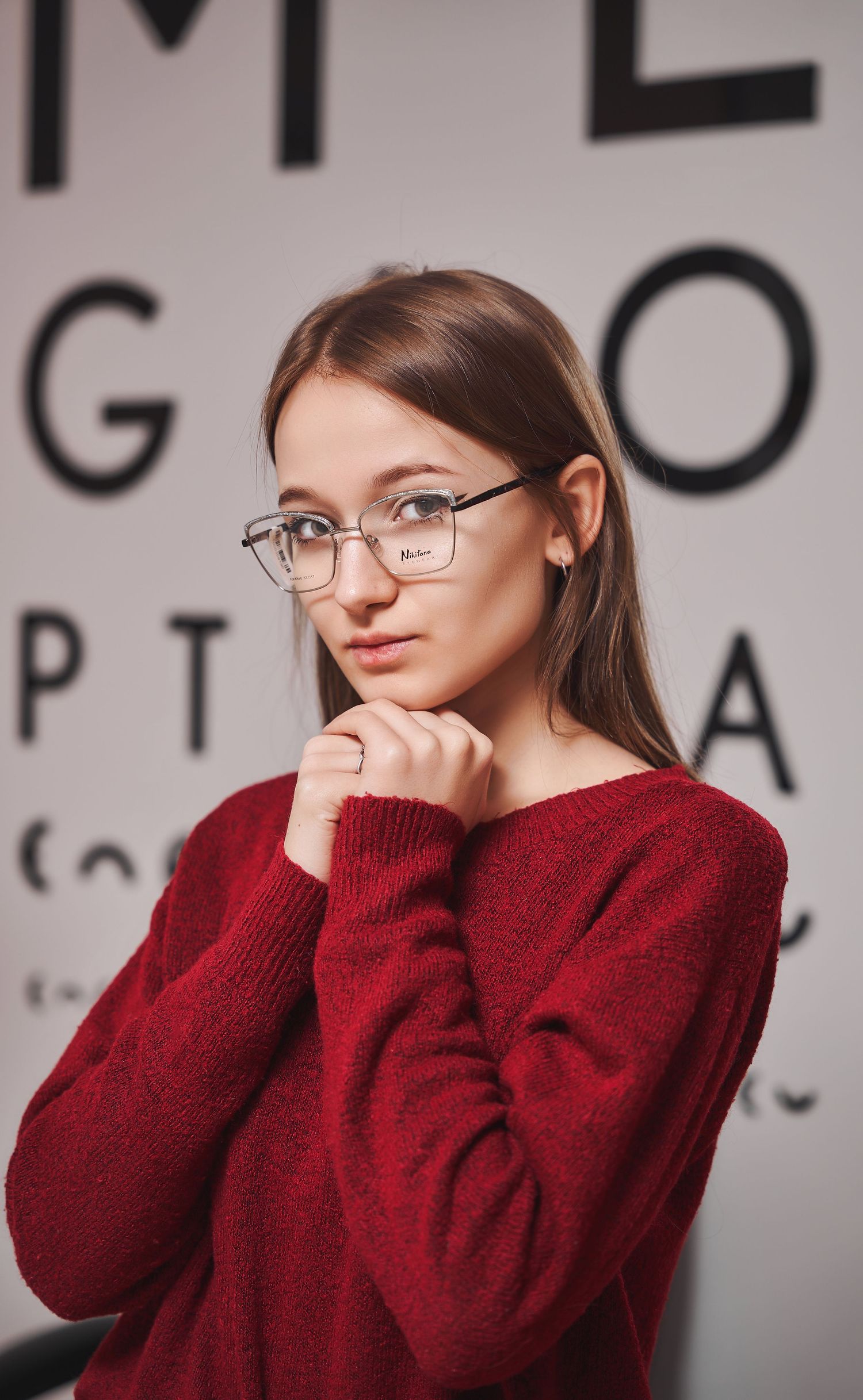 Person in burgundy sweater posing near eye chart in medical setting.