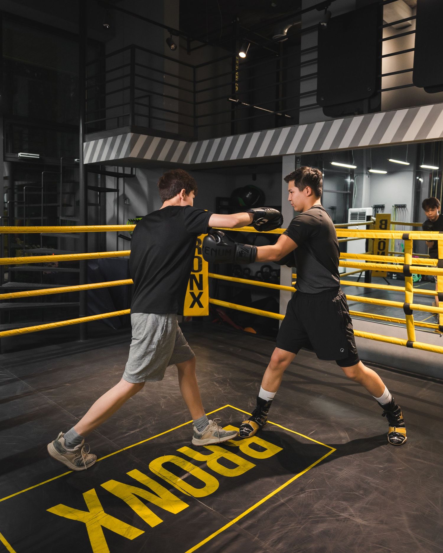 Two people practicing boxing footwork drills on marked floor with yellow barriers.