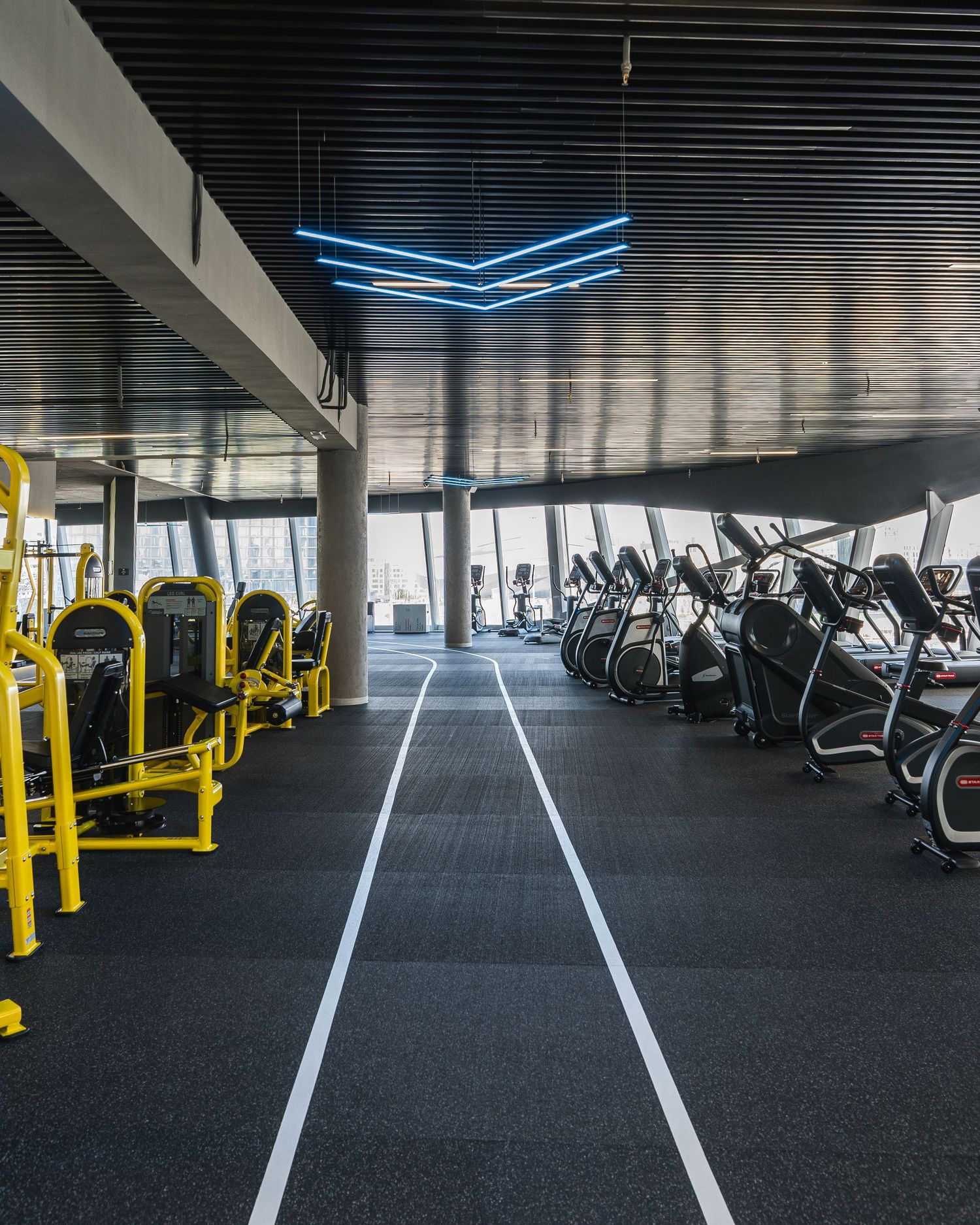Large fitness center with yellow exercise equipment and white floor markings in an industrial space.