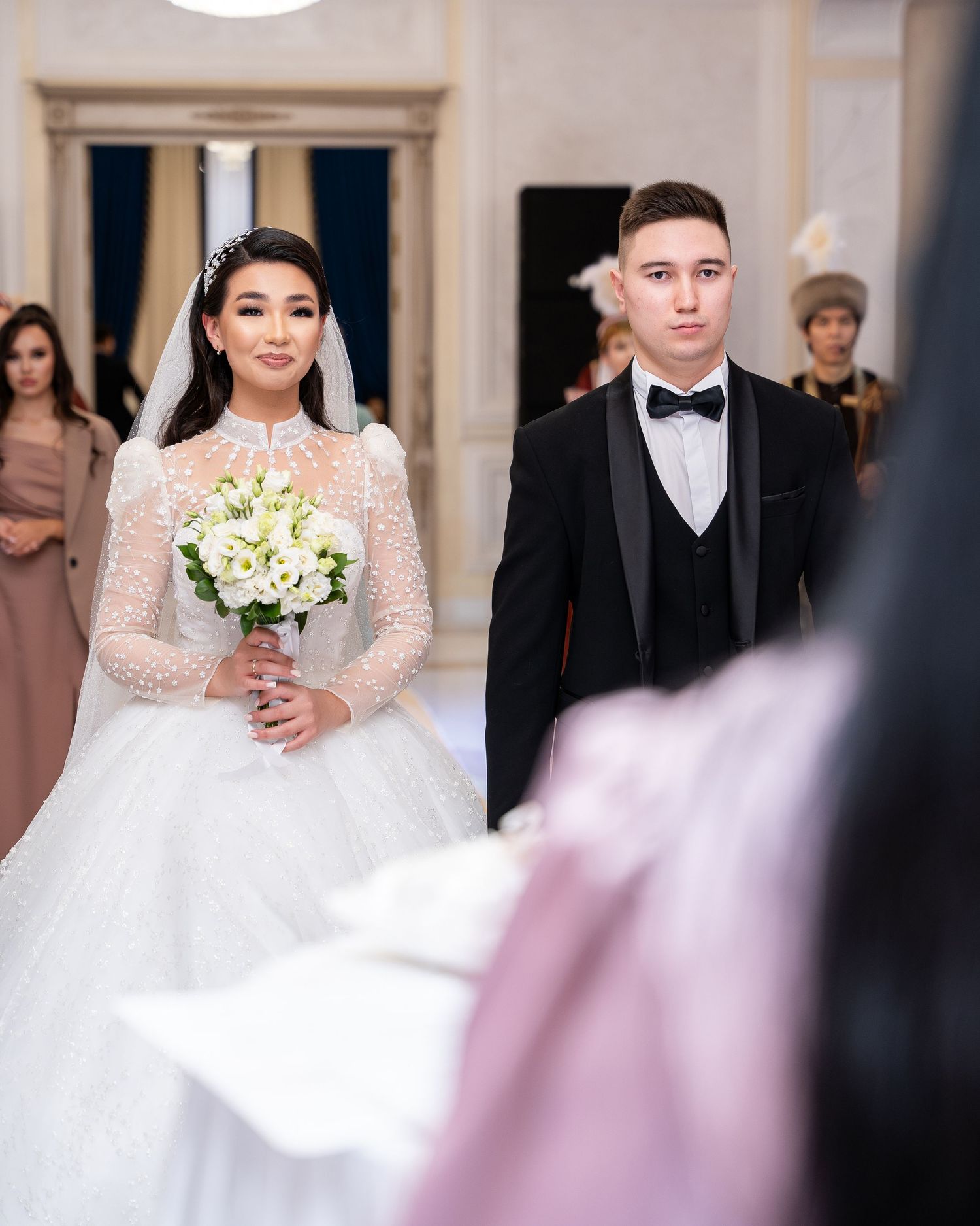 Bride in stunning white ballgown with veil holds bouquet standing beside groom in black tuxedo at wedding.