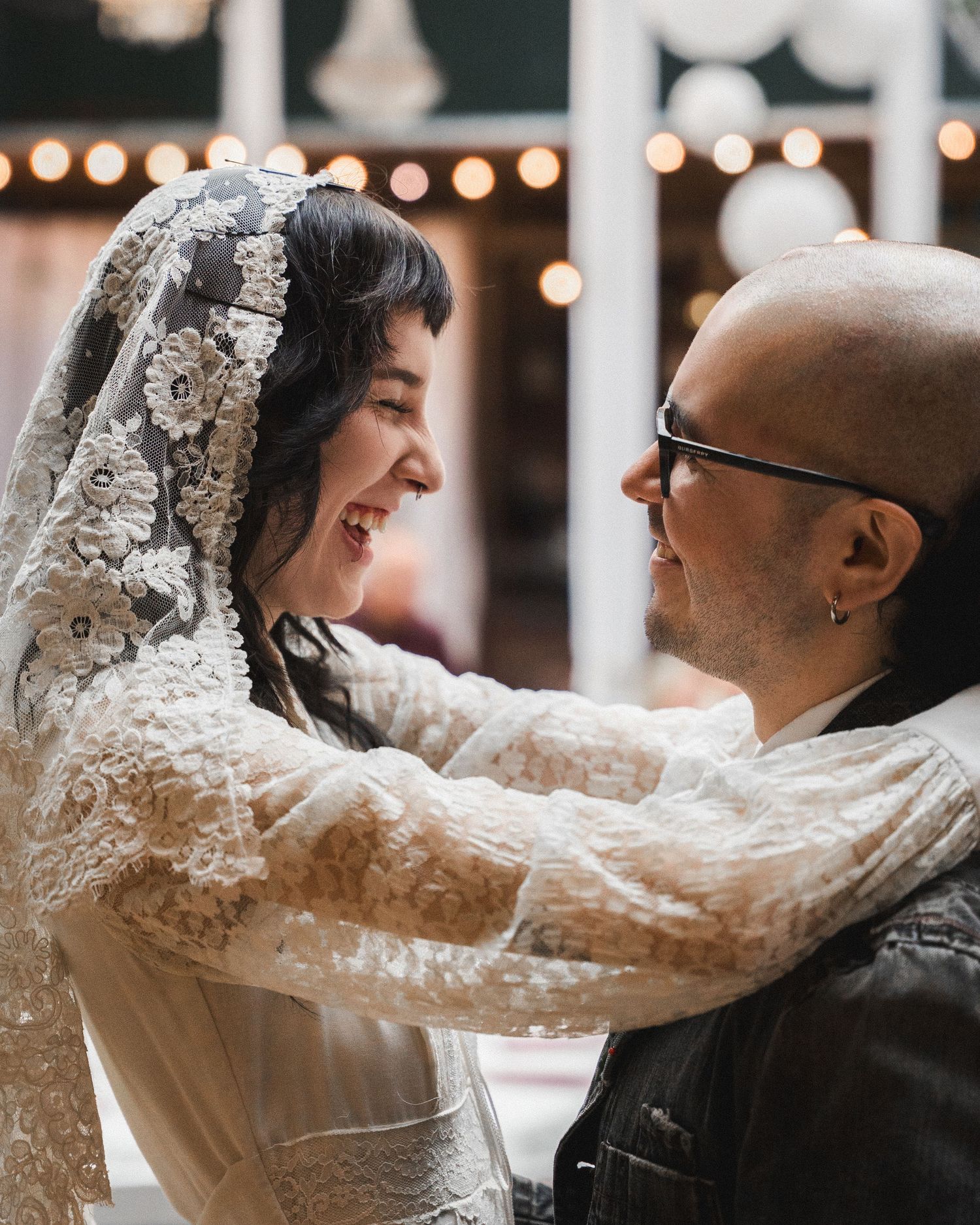 A joyful bride in lace veil and groom in black jacket share a tender moment at their wedding reception.