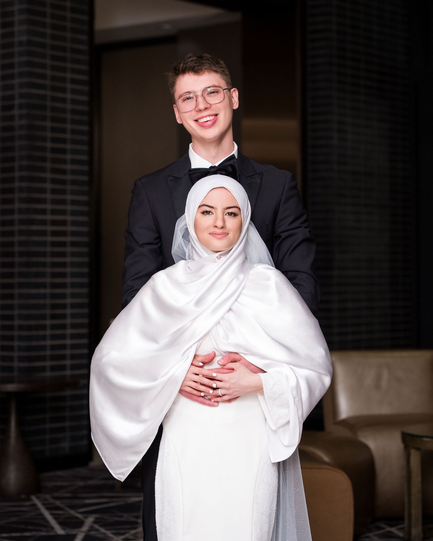 Couple in formal wedding attire, groom in tuxedo embracing bride wearing white hijab and gown, posing indoors.