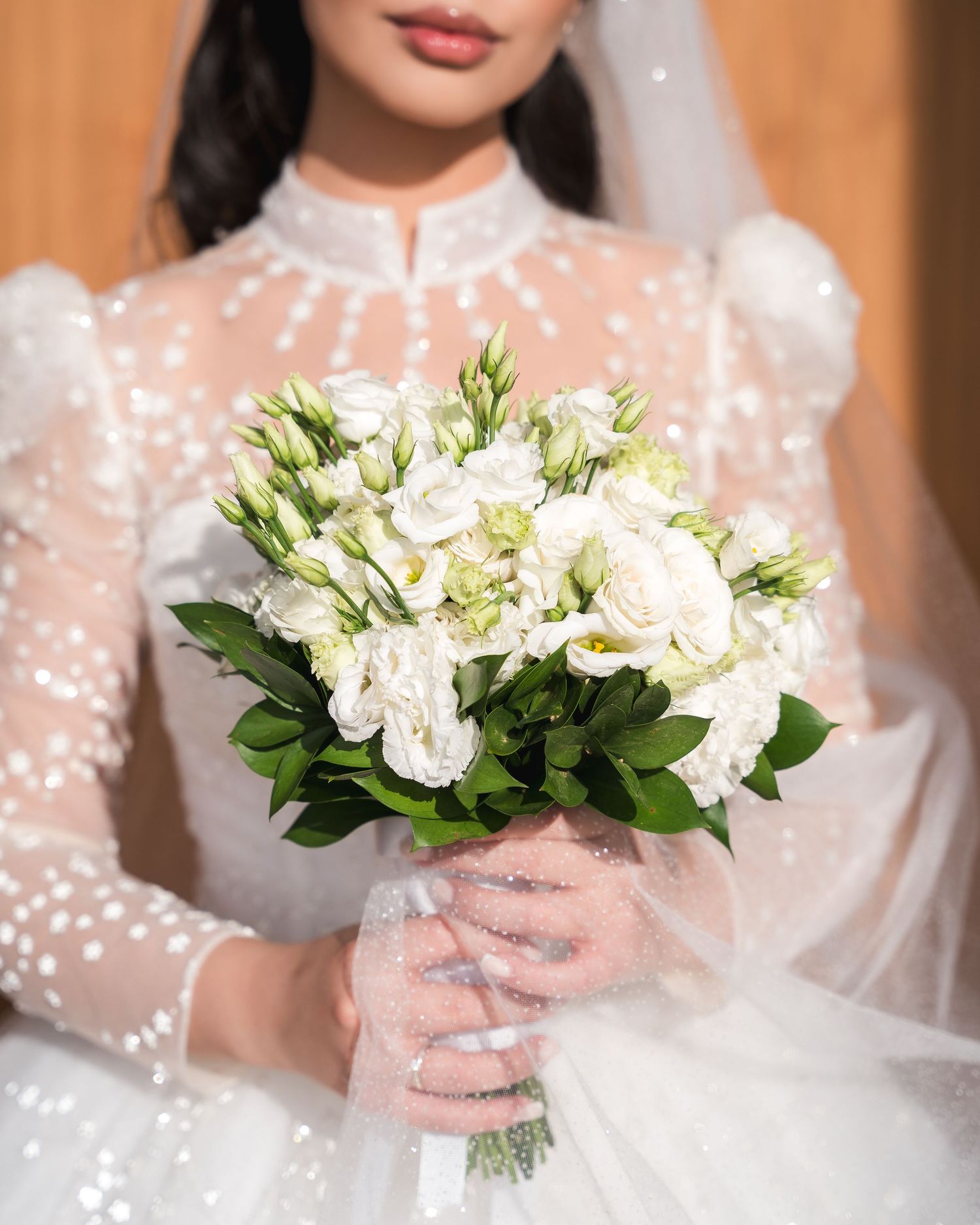 Close-up of bride in pearl-dotted sheer white wedding gown holding lush bouquet of white lisianthus flowers.