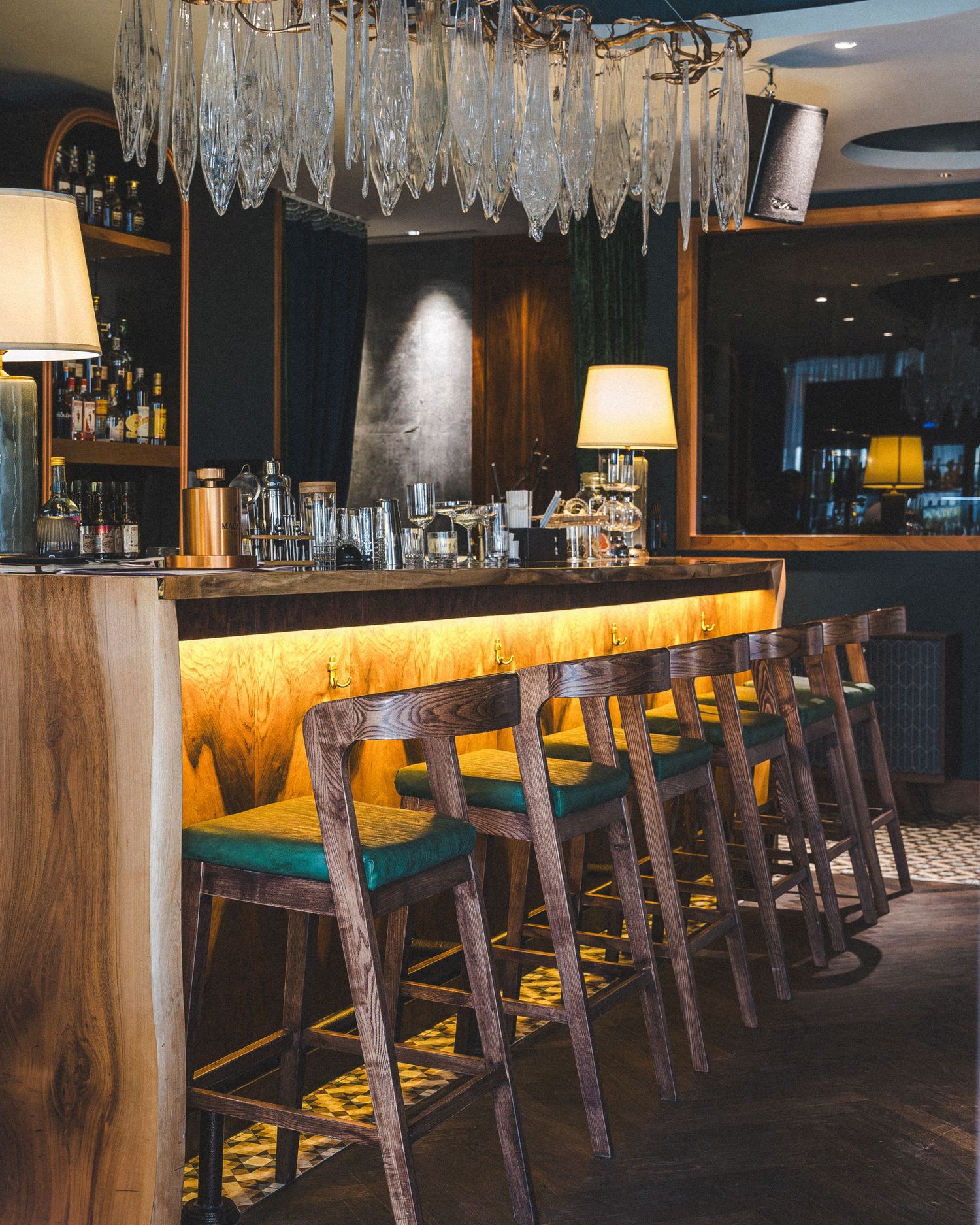 Modern bar with crystal chandelier, wooden counter, and turquoise upholstered bar stools.