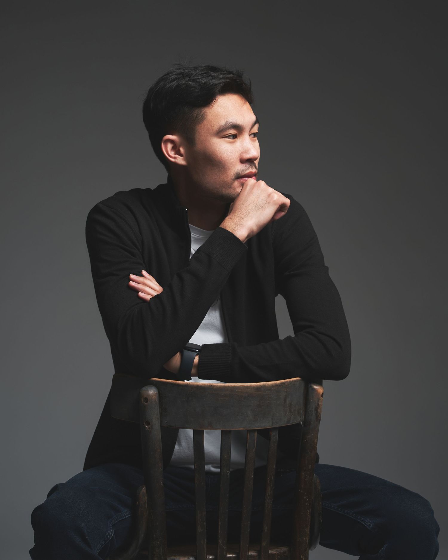 Person in black outfit sitting thoughtfully on wooden chair against dark background.