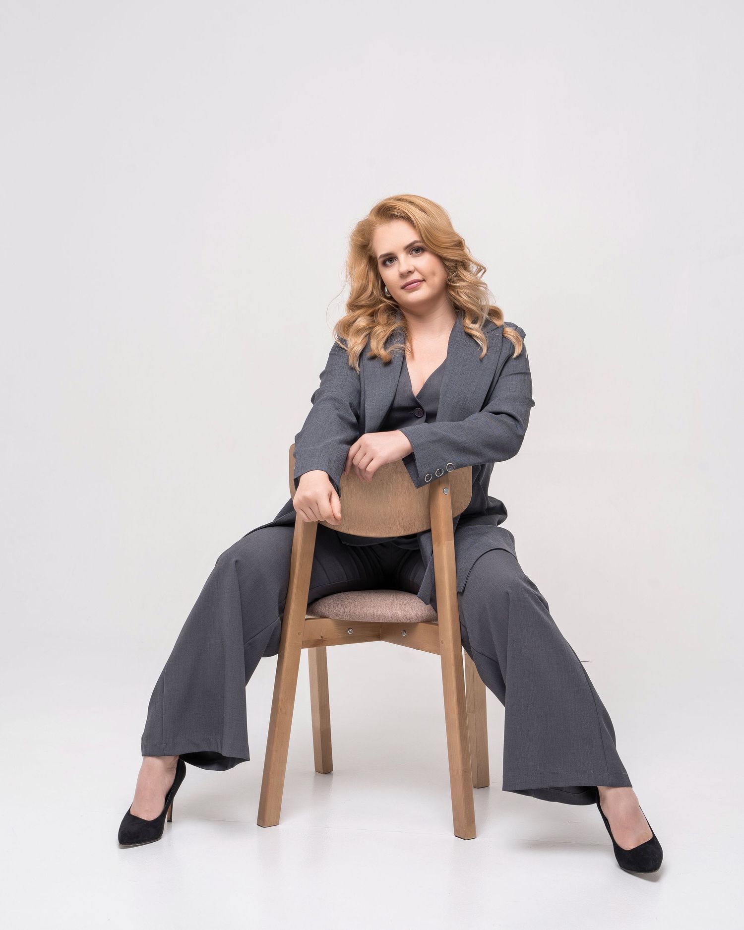 Woman in gray suit poses confidently on wooden chair in studio setting.