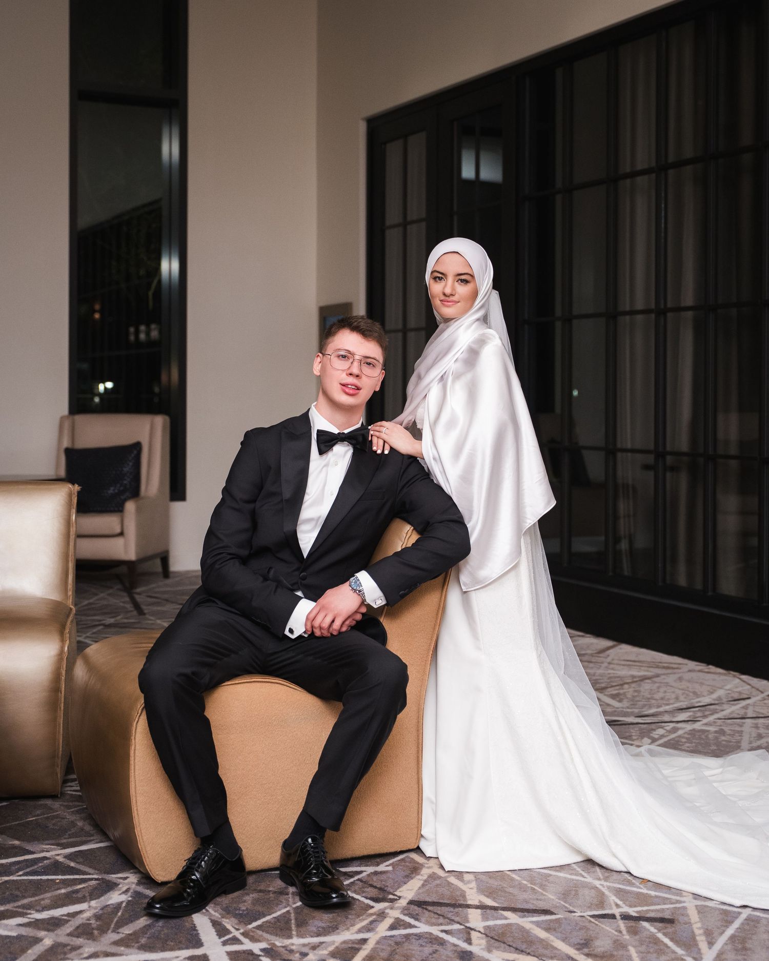 Elegant wedding couple posing indoors, groom in black tuxedo seated, bride standing in white gown and hijab.