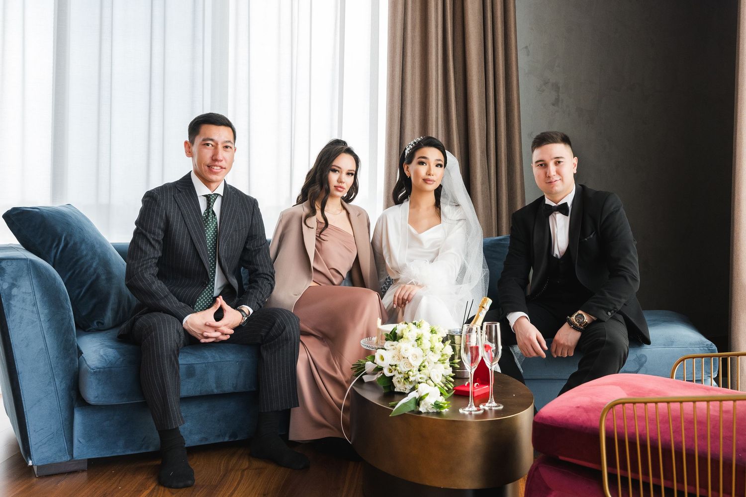 Wedding party of four posing on blue sofa, bride in white gown with bouquet and champagne on table.