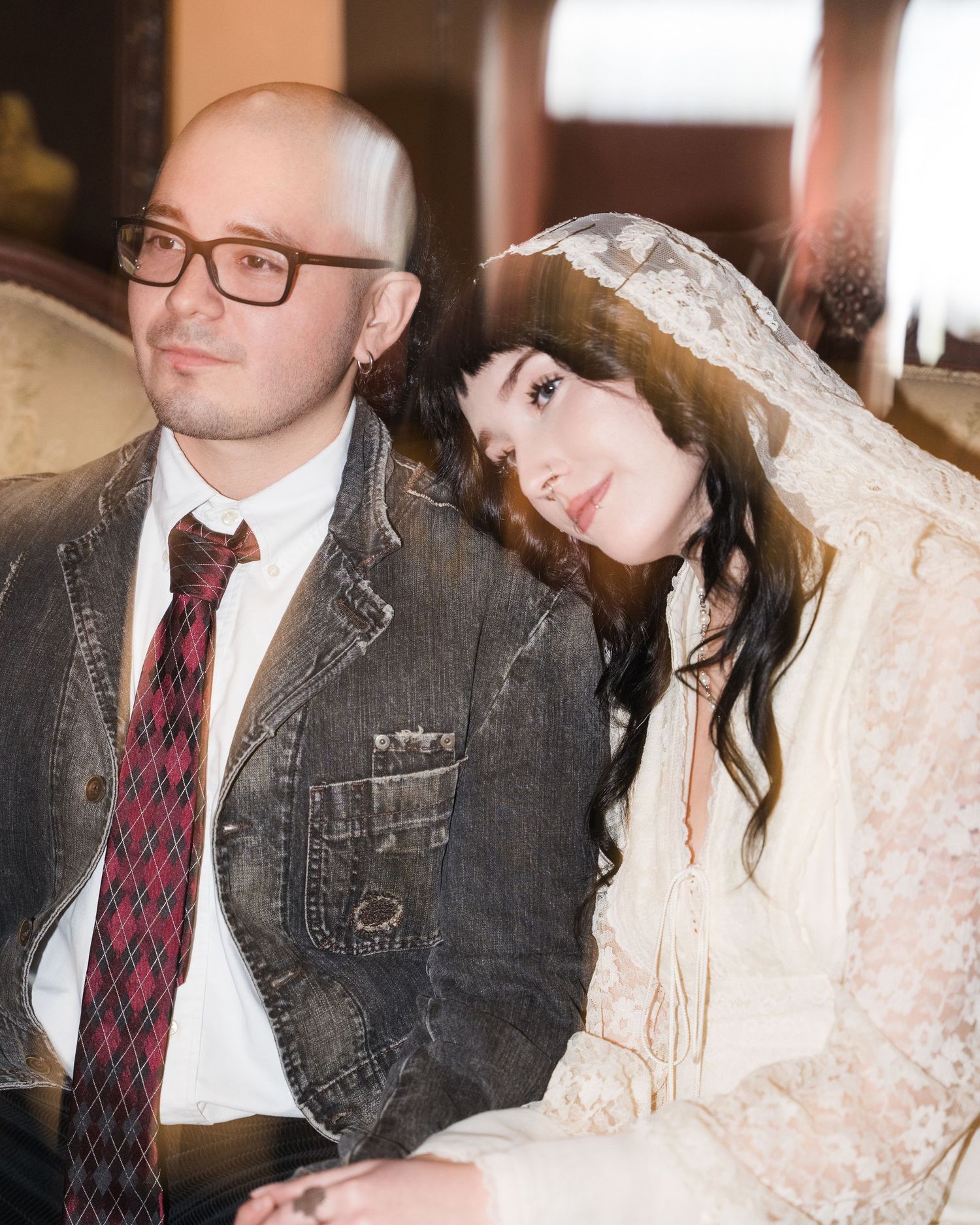 Bride in vintage lace veil rests head on groom's shoulder in denim jacket and plaid tie at wedding.