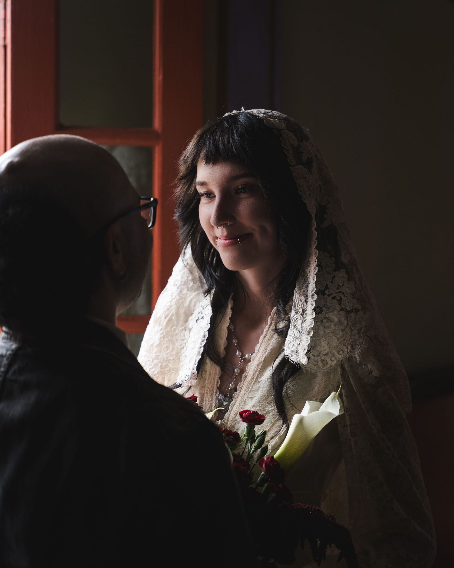 A smiling bride in a lace veil holds a bouquet of roses and calla lilies while facing a bald man in glasses.