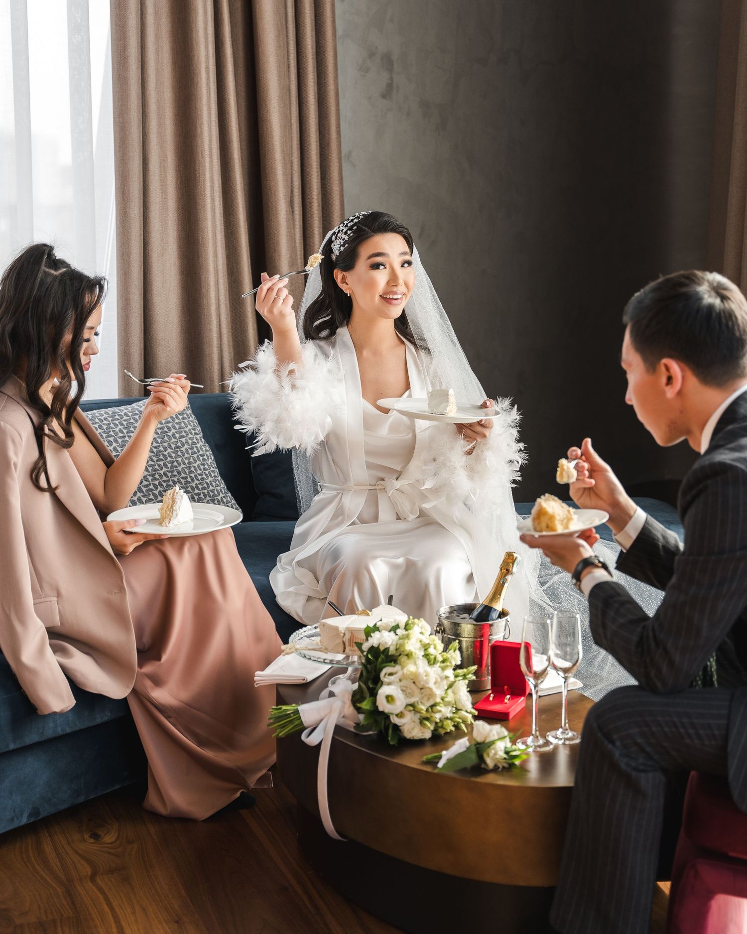 Bride in white feather robe celebrates with bridesmaids and groom over cake and champagne.