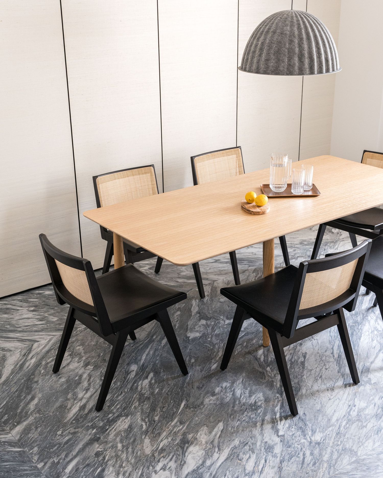 Minimalist dining setup featuring light wood table with angular black chairs on grey marble floors.