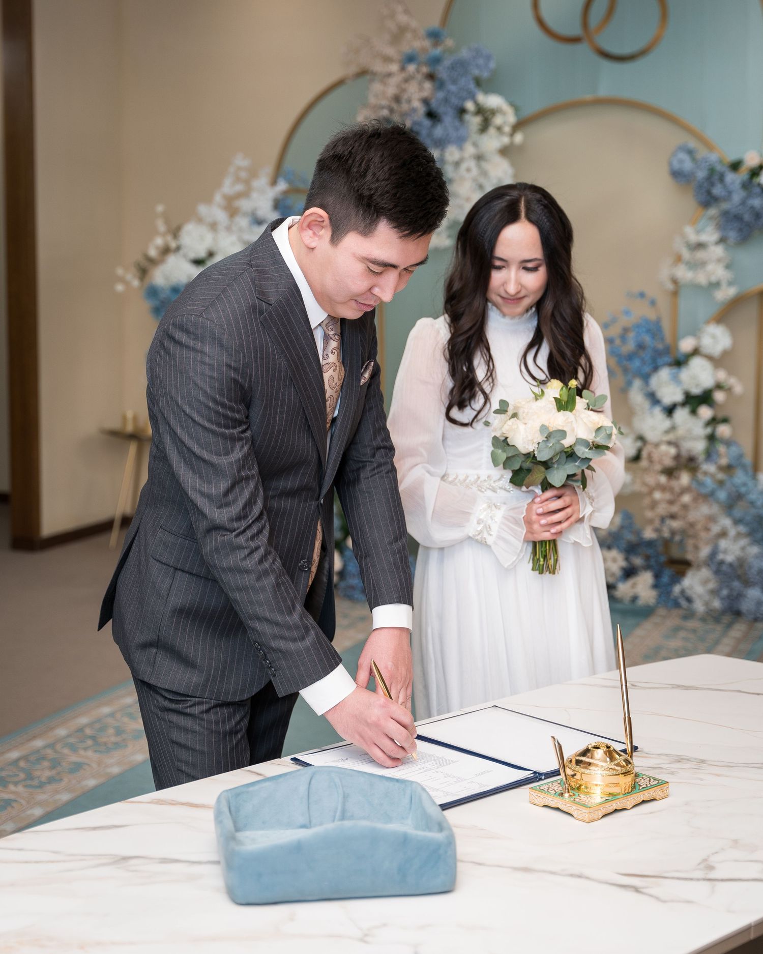 Groom signing marriage certificate at registry office while bride in white dress holds flower bouquet.