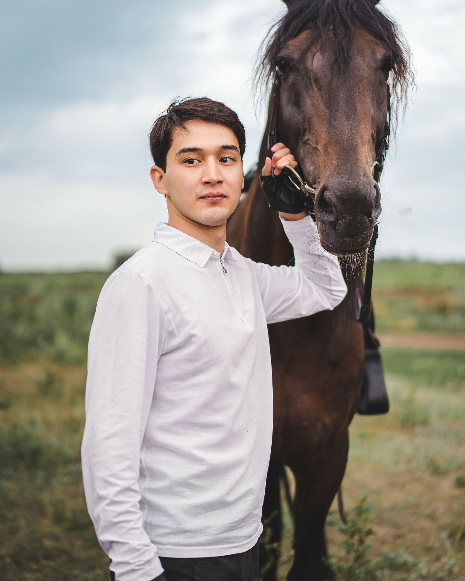 Person in white shirt posing with dark brown horse in outdoor field setting.