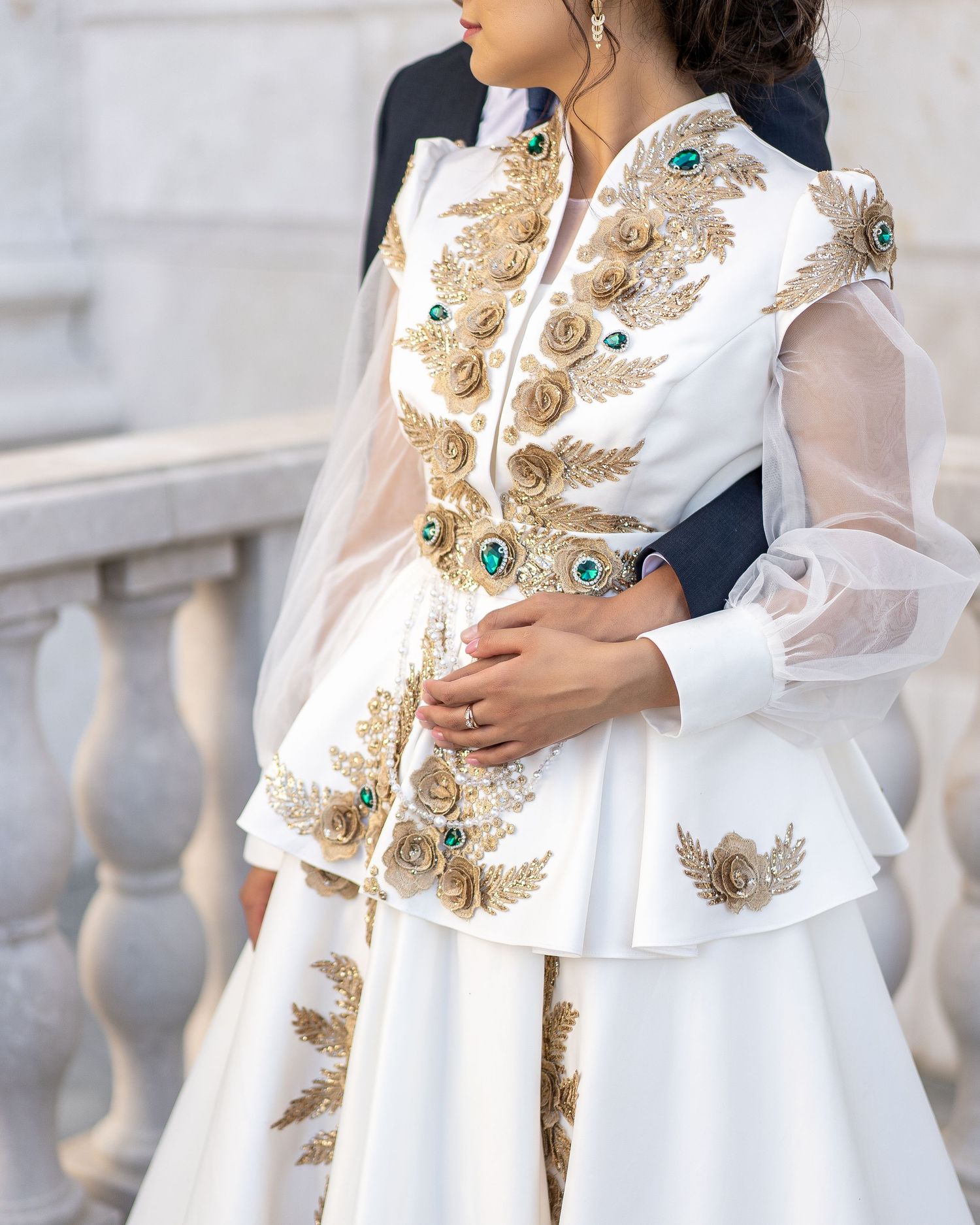 White peplum wedding dress with gold embroidery and emerald gems, featuring sheer organza sleeves.