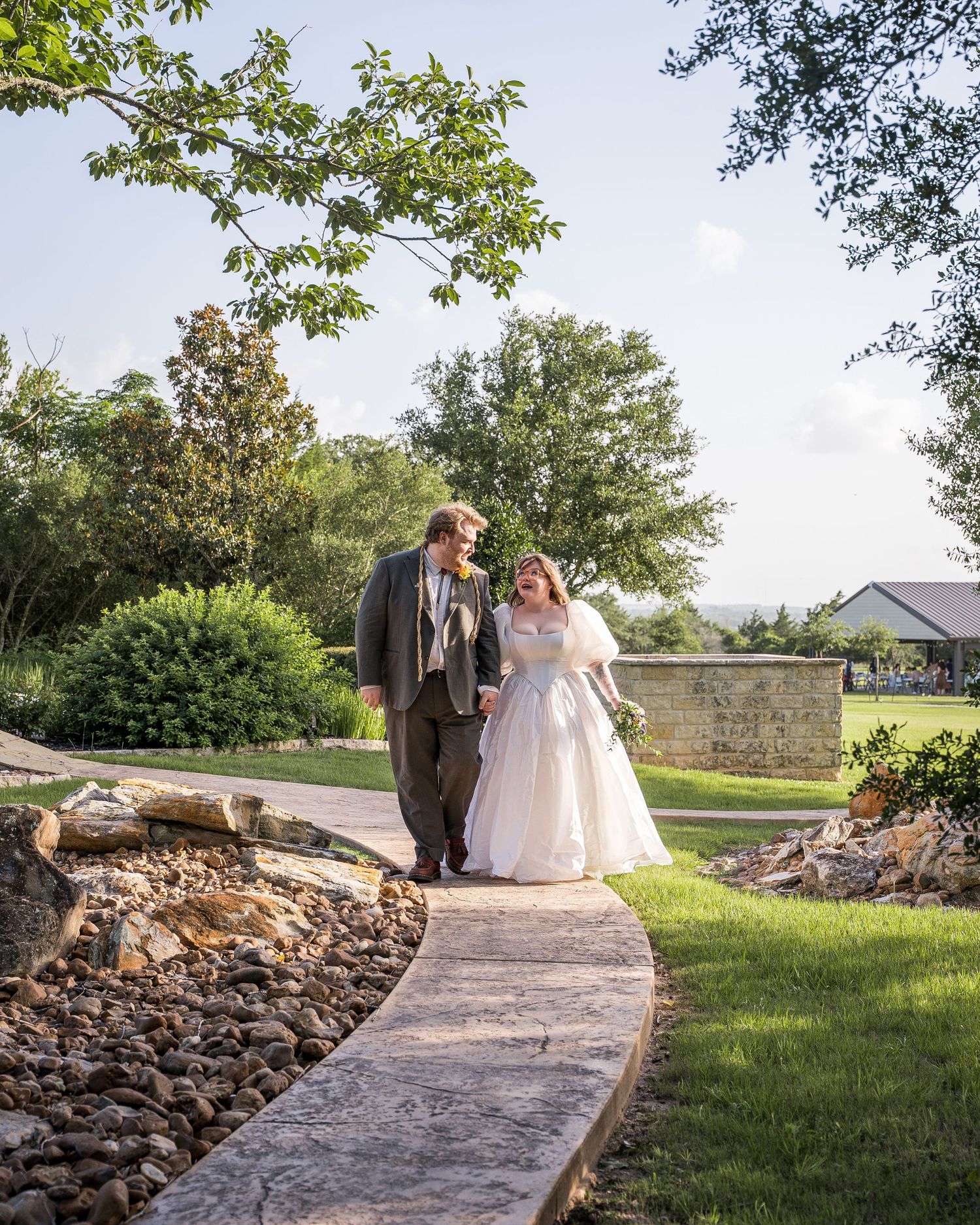 Bride and groom walking hand in hand along a stone path in a lush garden wedding venue.