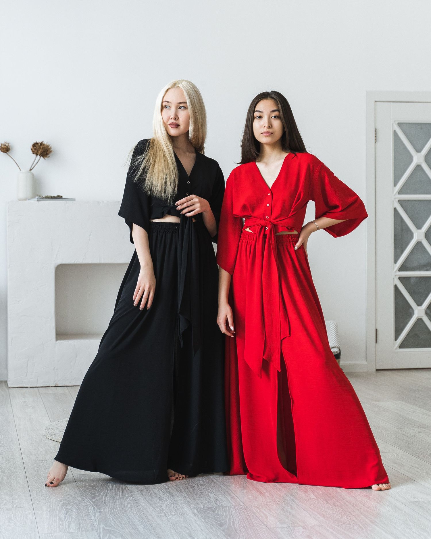 Fashion editorial with models in flowing black and red maxi dresses posed together on light floor.