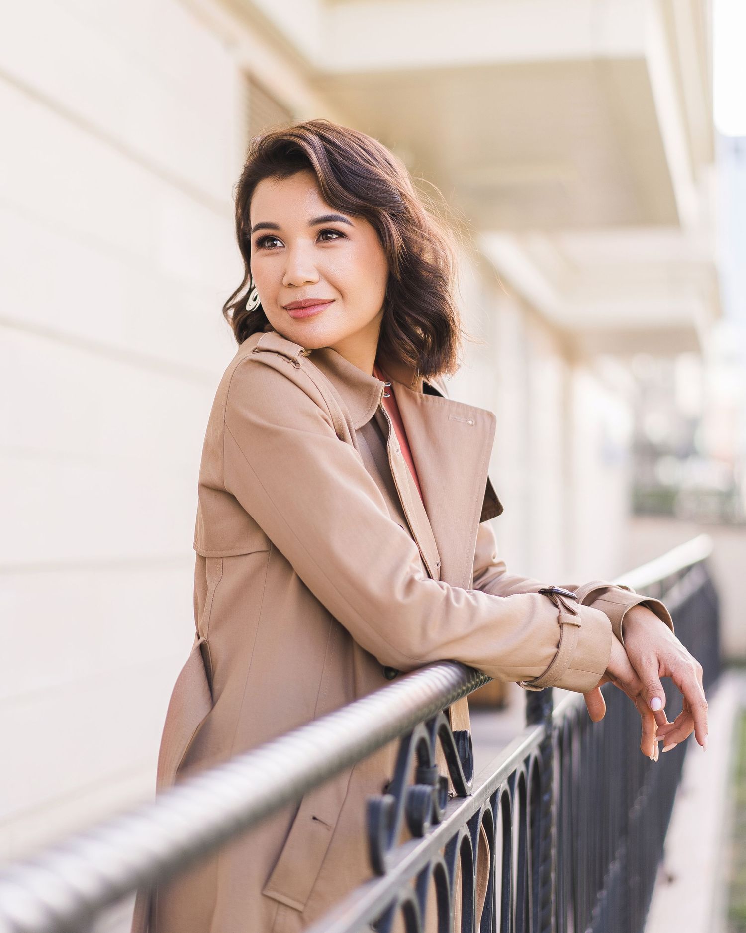 Person in tan blazer leaning on metal railing outdoors with modern architecture backdrop.