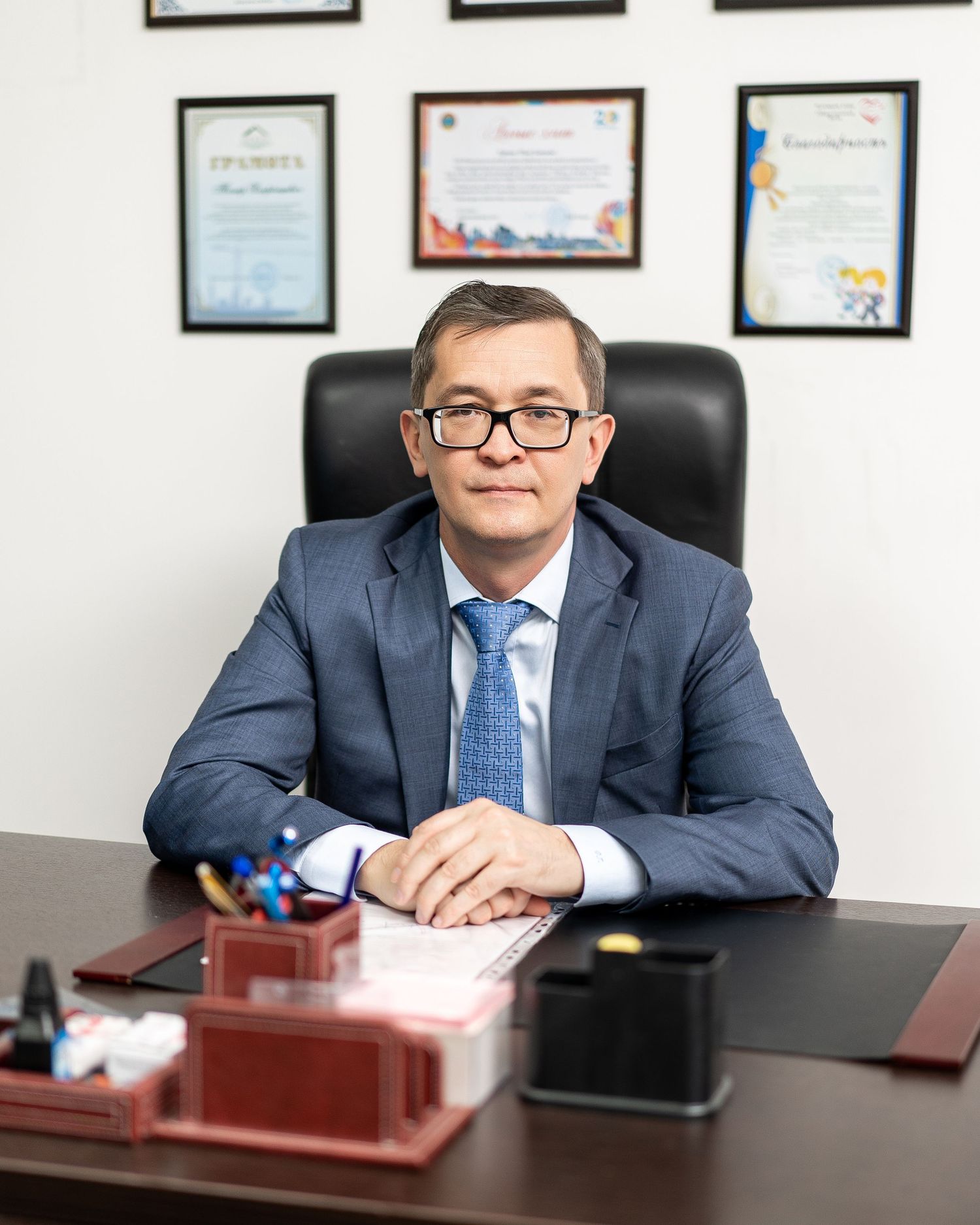 Executive seated at desk with certificates and awards displayed on wall behind.