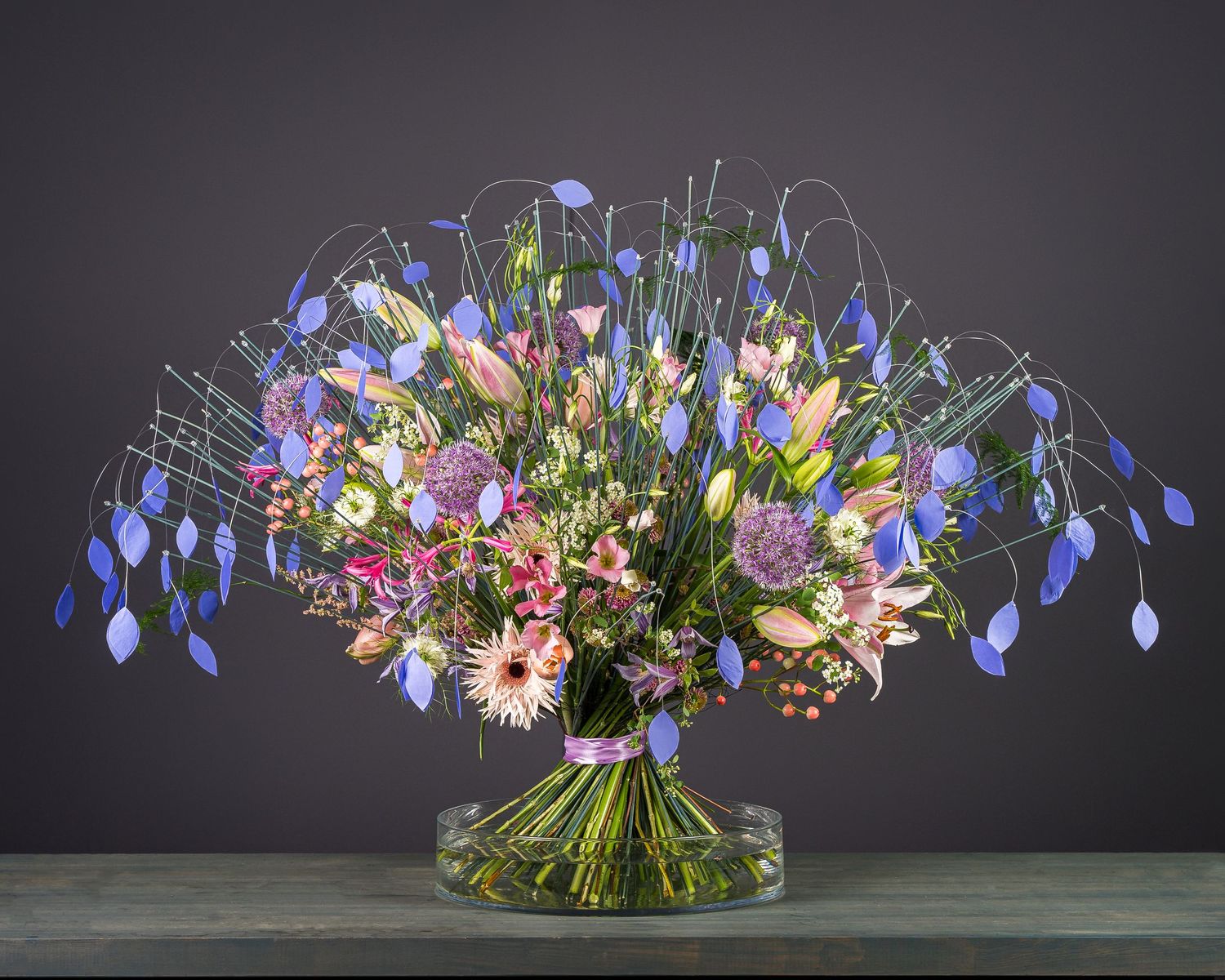 Vibrant floral arrangement with blue delphiniums and pink lilies in clear glass vase.