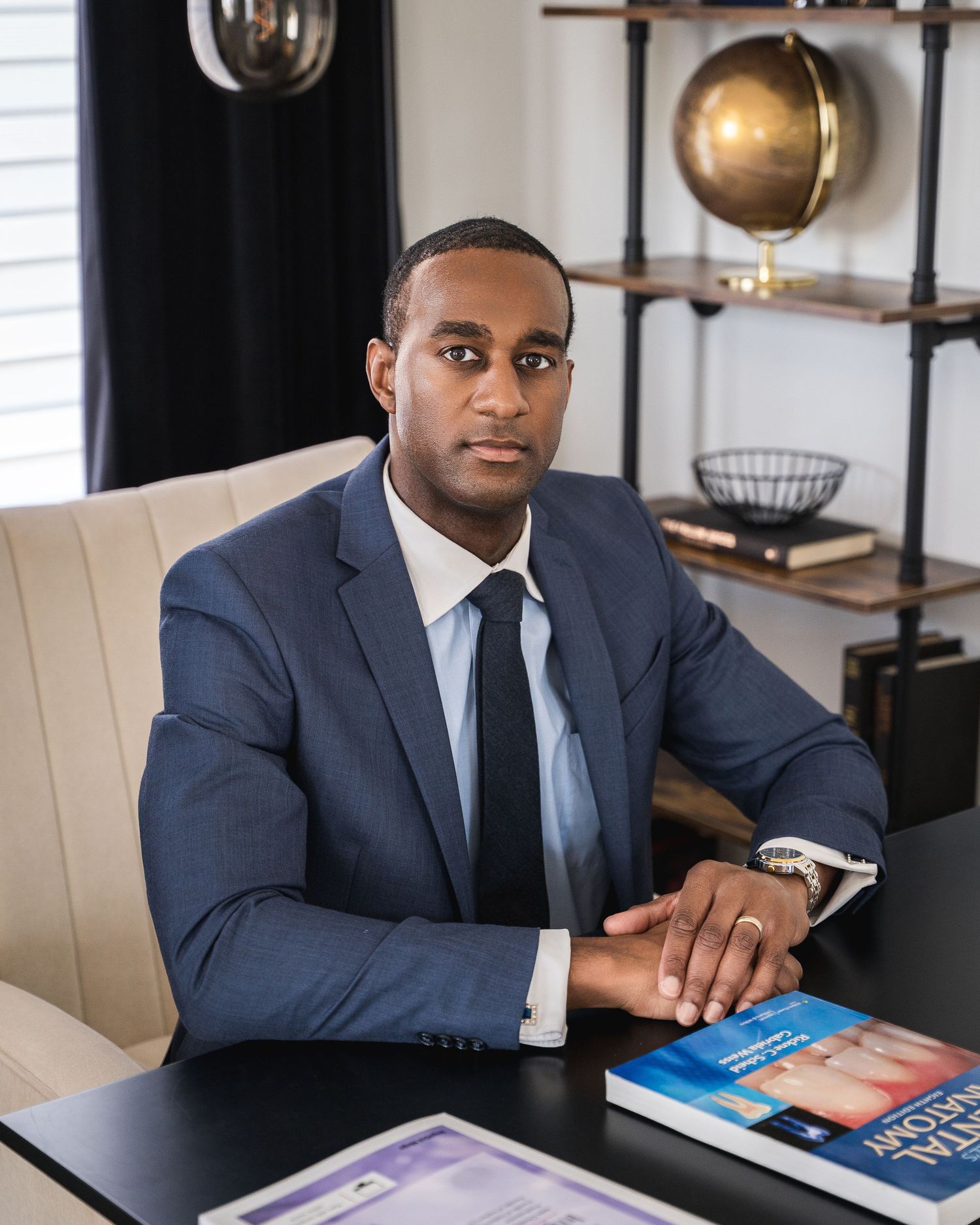 Professional in navy suit sitting at desk with books in modern office setting.