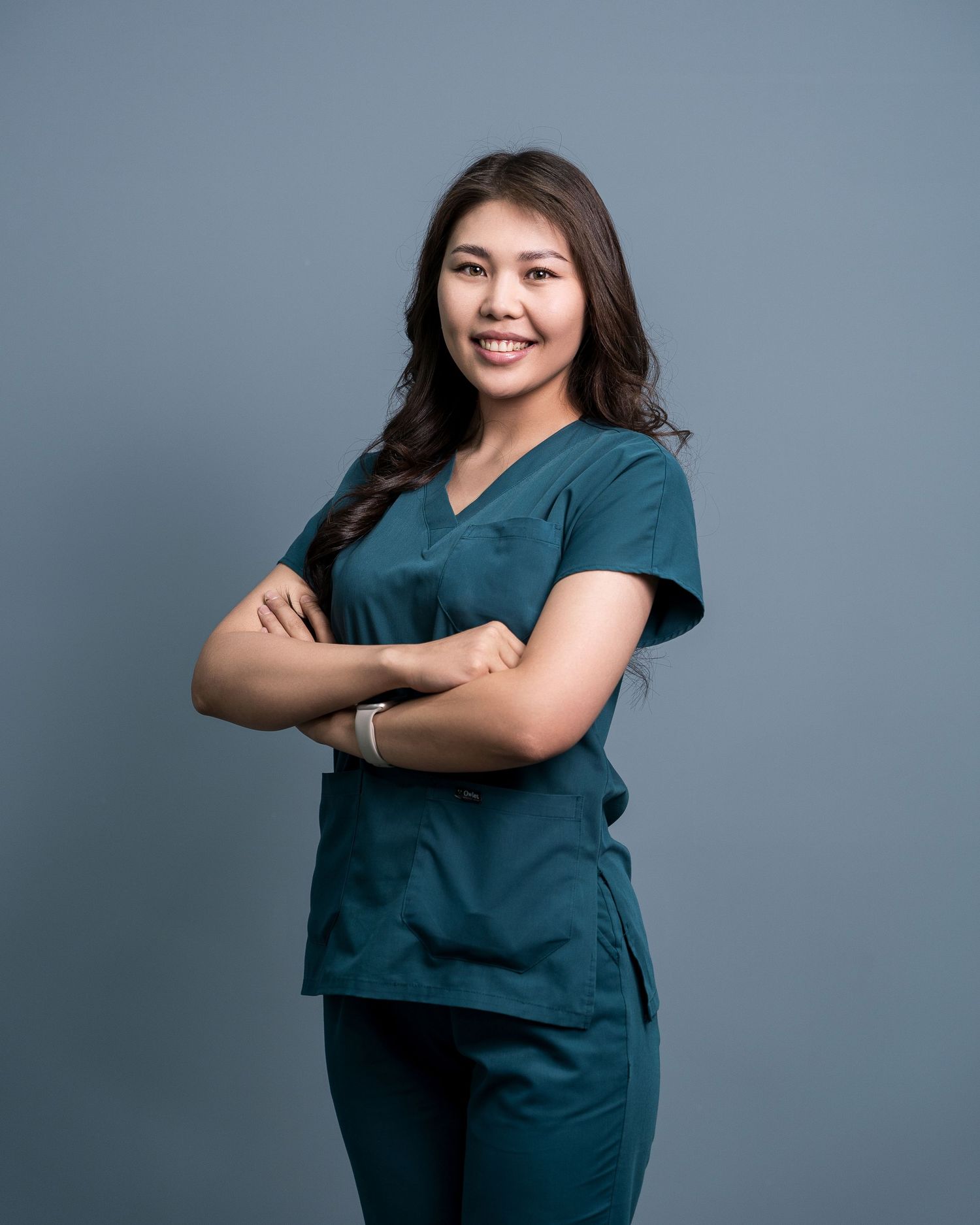 Healthcare worker in teal scrubs poses confidently with crossed arms against gray background.