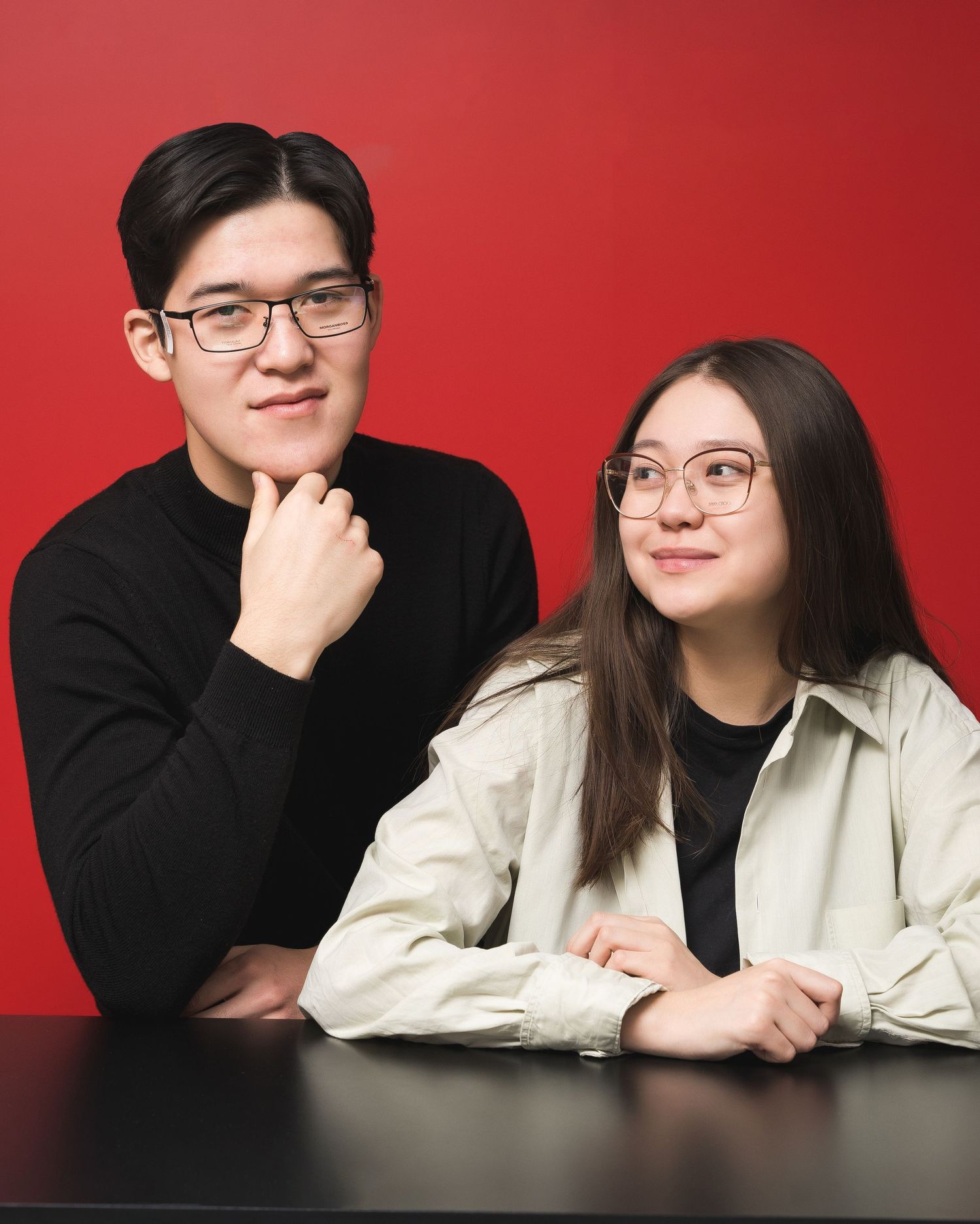 Couple in black and white outfits posing together against red background.