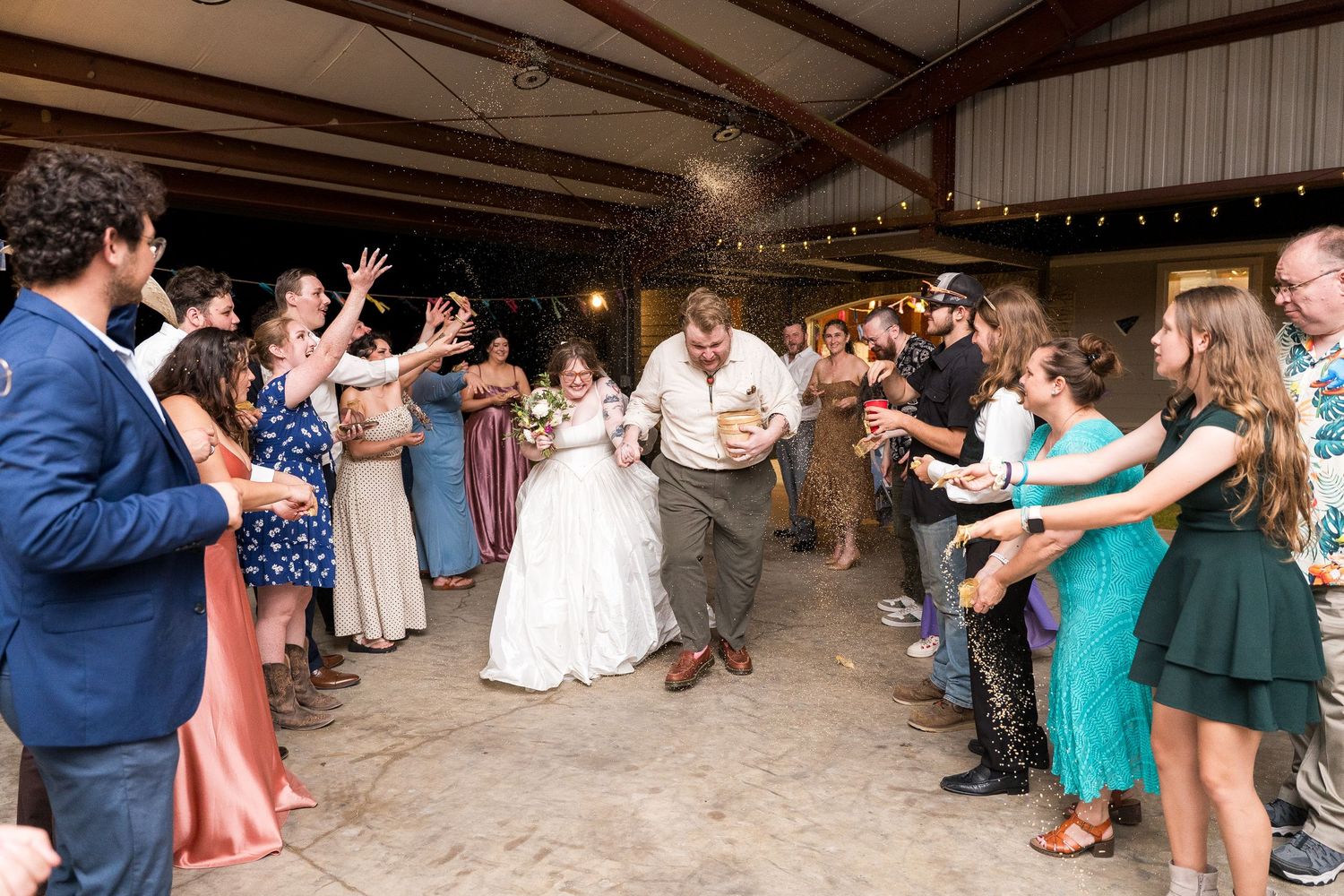 Bride and groom exit wedding reception through cheering guests tossing rice in rustic barn venue.